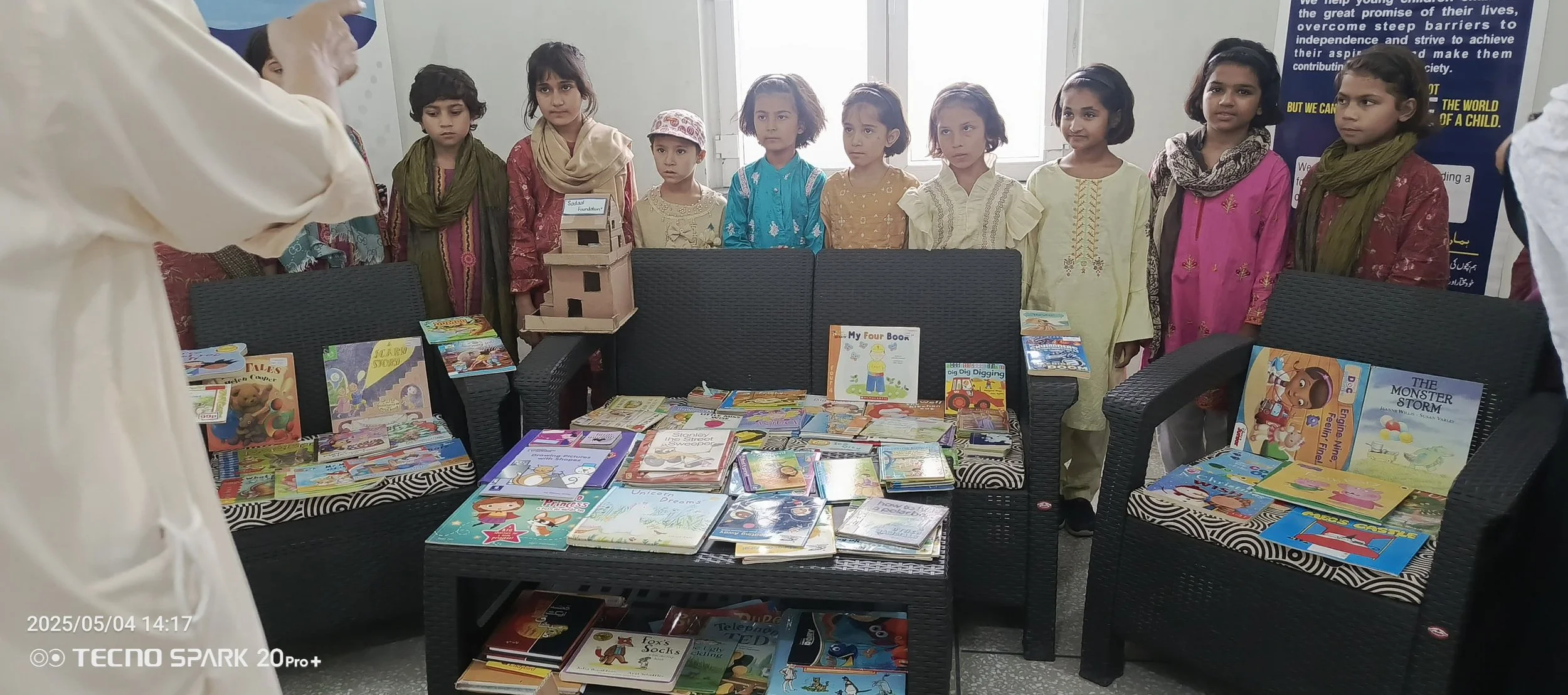 A group of children, mostly girls, standing behind a display of children's books in a room with large windows. The children are wearing colorful traditional dresses and looking at an adult who is talking to them.
