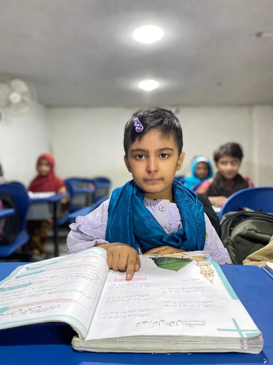 A young boy sitting at a school desk in a classroom, pointing to an open textbook, with other children seated behind him.