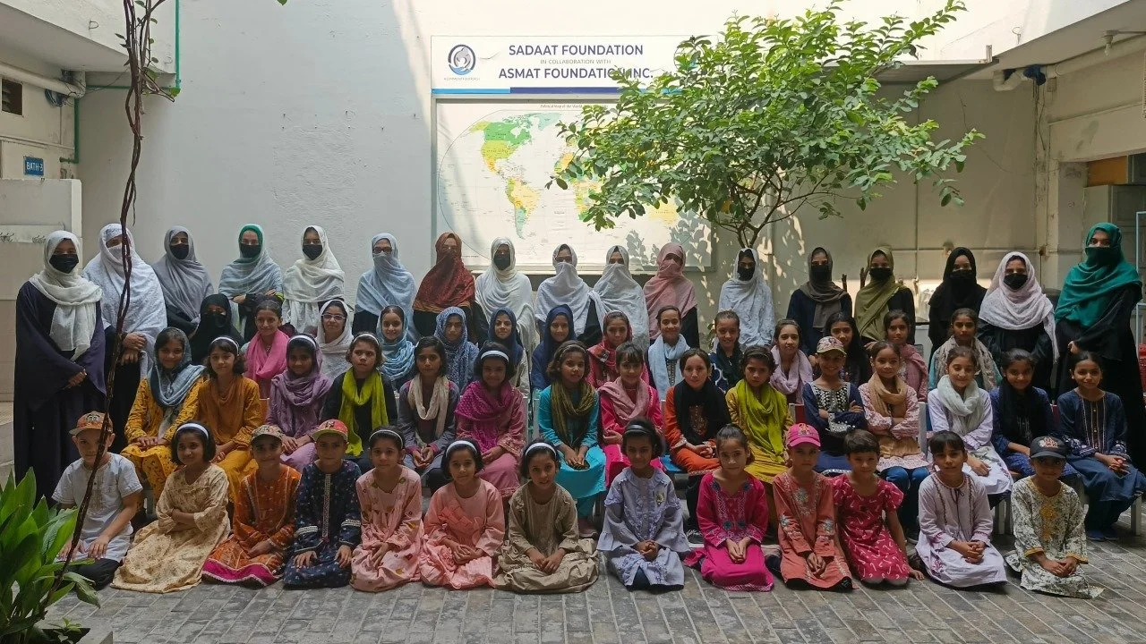 Group of school children and teachers posing for a photo outside, with a map of the world behind them, in a courtyard with white walls and a tree.