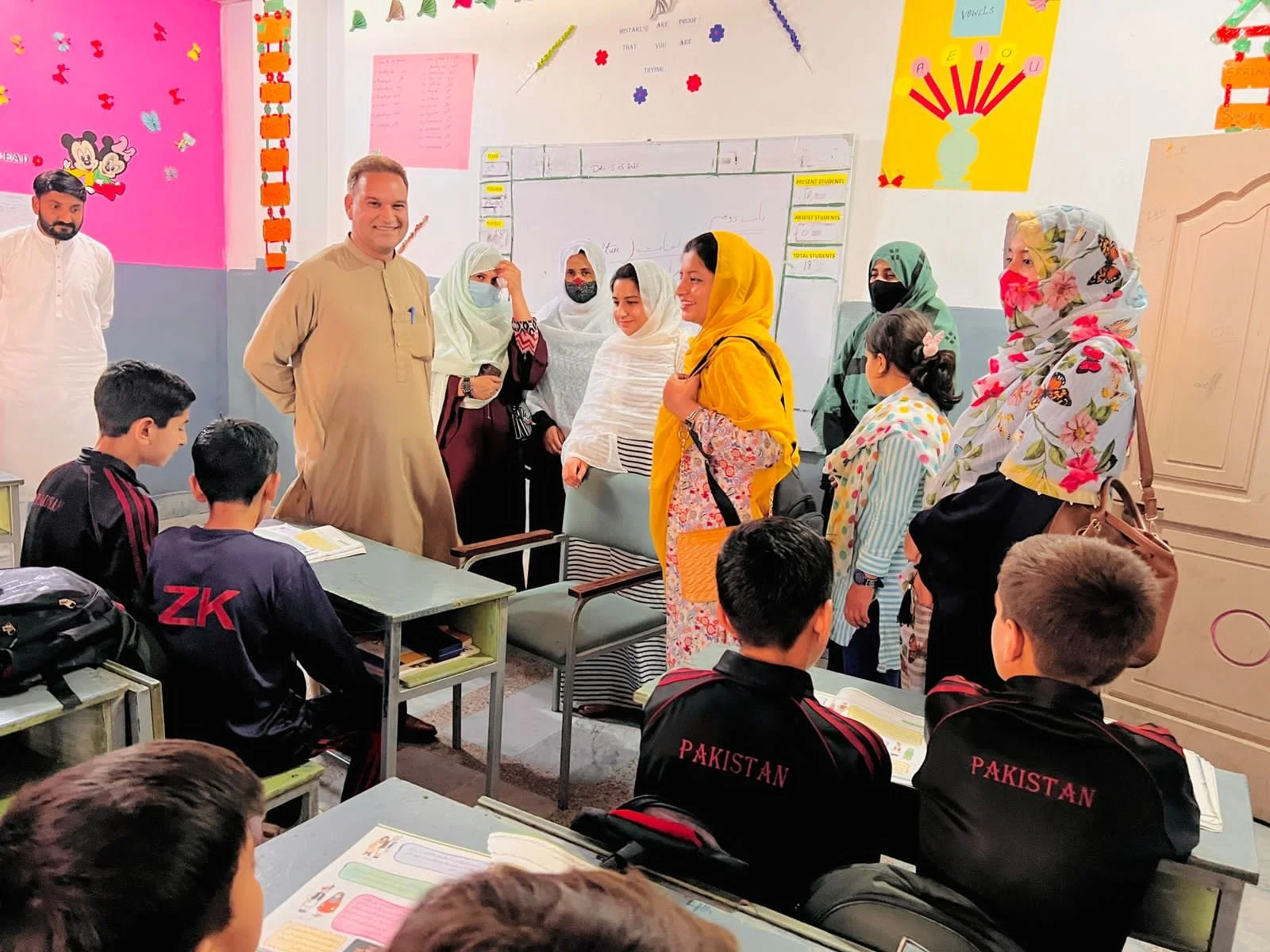 A classroom scene with students of Pakistani origin seated at desks, and teachers or visitors, mostly women wearing colorful headscarves, standing at the front of the room. Some students are reading their textbooks, and the classroom walls are decorated with educational posters and colorful artwork.