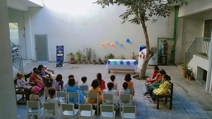 Children seated in a courtyard facing a table decorated with balloons, with a potted plant and a tree next to the table.