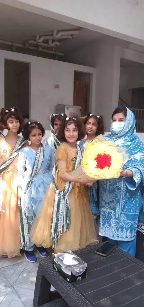 A group of young girls in traditional party dresses standing in a line being presented with a yellow flower bouquet by a woman in a blue shawl and face mask.