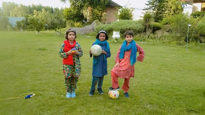 Three young girls standing on a grassy field outdoors, each holding a soccer ball. One girl is wearing a floral dress and blue shoes, another is in a blue traditional dress with a headband, and the third girl is dressed in a pinkish traditional outfit with a blue scarf. Trees and a house are visible in the background.