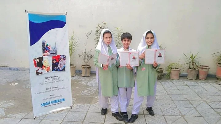 Three children in green and white uniforms holding open books, standing outside next to a banner with images of children and contact information, in front of potted plants.