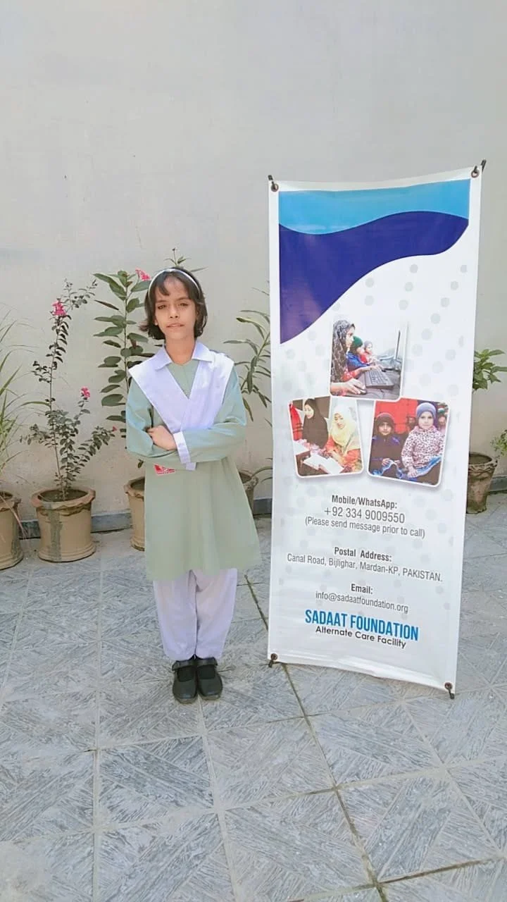 A young girl in a school uniform standing next to a banner for Sadaat Foundation, with potted plants in the background.
