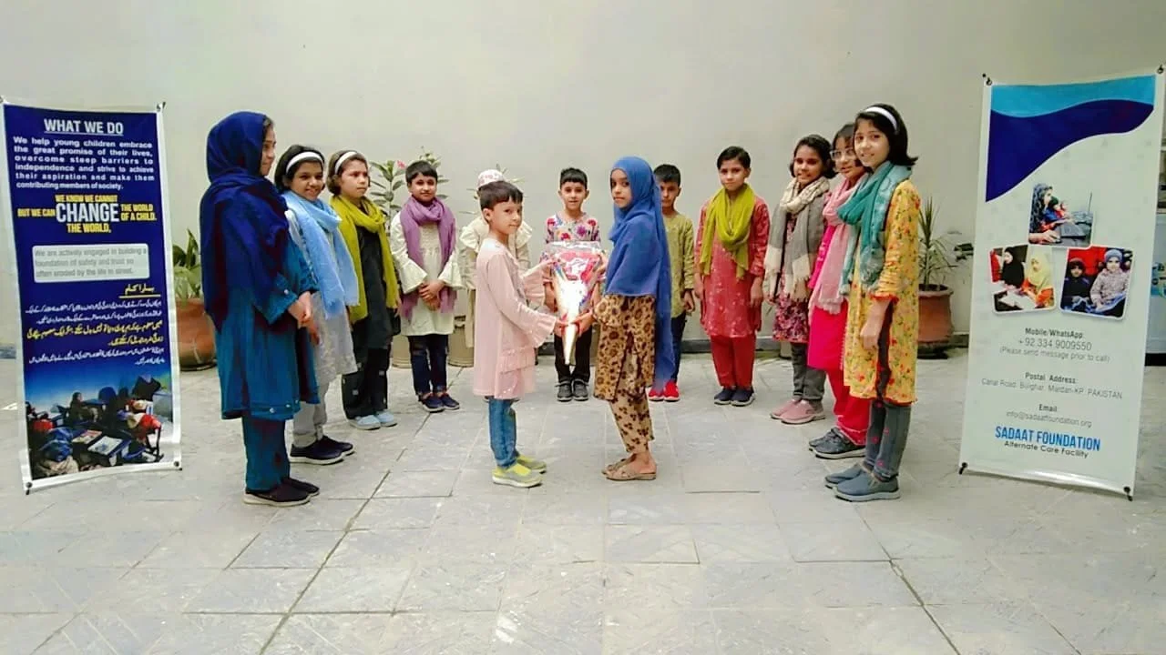 Children participating in an indoor group activity, with two girls at the center exchanging a gift or award, flanked by posters with informational and organizational details.
