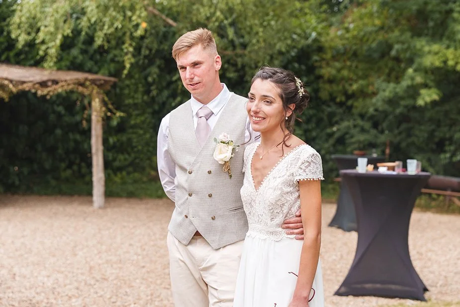 Un couple lors d'un mariage dehors, la femme porte une robe blanche et l'homme une veste beige avec cravate. Ils sourient et posent ensemble.