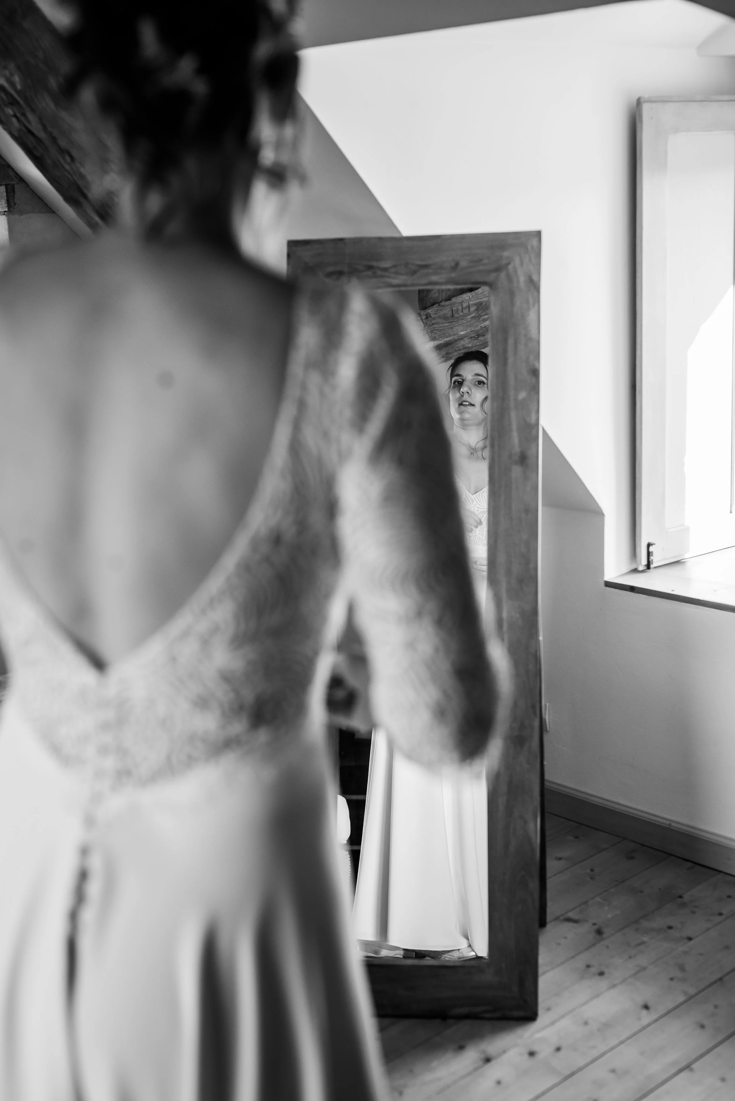 Une femme regarde son reflet dans un miroir en bois, vue de derrière, dans une pièce avec sol en bois et fenêtre. La photo est en noir et blanc.