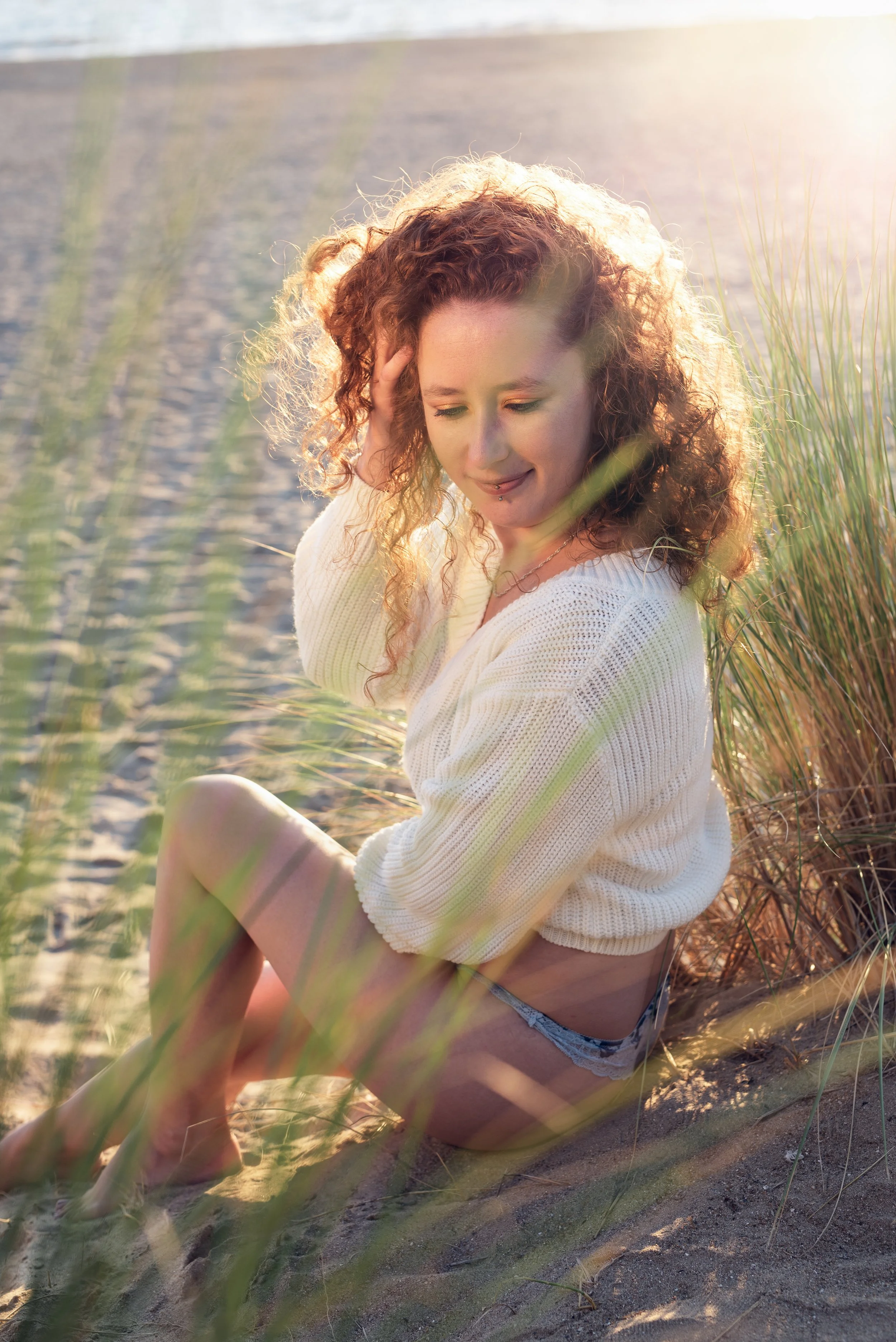 Une jeune femme aux cheveux bouclés roux assise sur la plage, portant un pull blanc et un short en denim, souriant doucement, dans un décor ensoleillé avec des herbes hautes autour d'elle.
