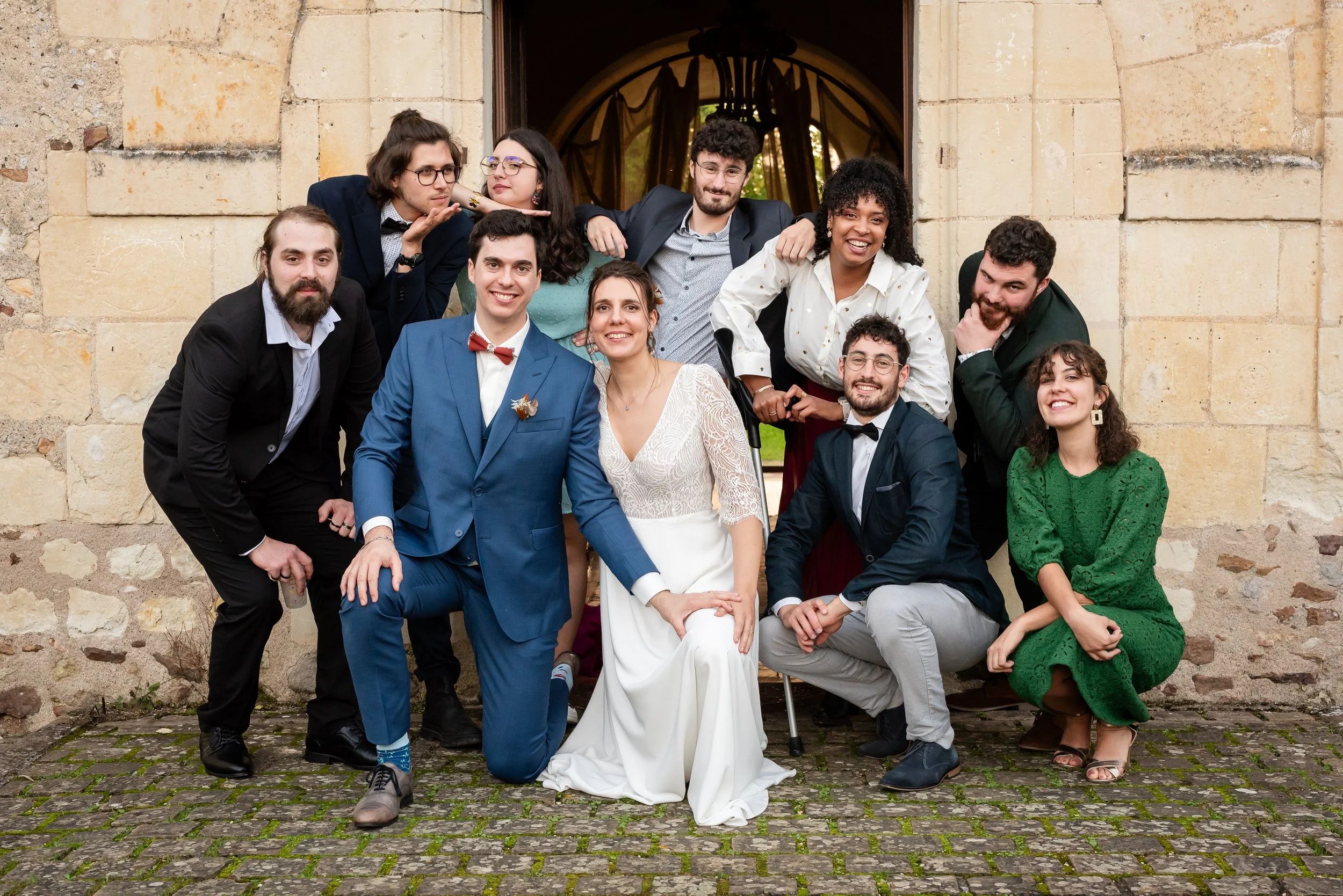 Groupe de personnes posant devant un ancien bâtiment en pierre, certains souriant, d'autres faisant des grimaces, avec des tenues variées, incluant un costume bleu avec un nœud papillon rouge, une robe blanche, des vêtements décontractés et élégants.