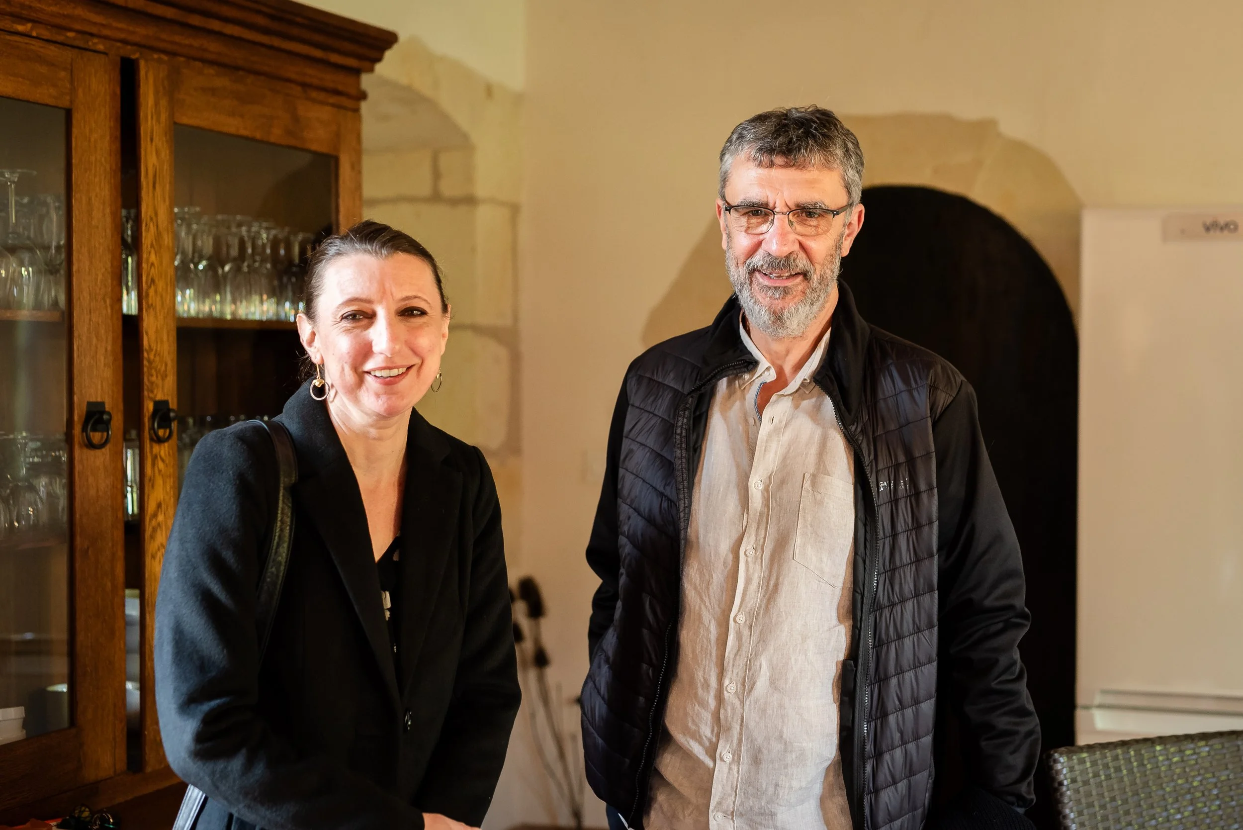 Deux personnes, une femme et un homme, posent ensemble dans une pièce chaleureuse. La femme porte un blazer noir, l'homme porte une chemise beige et une veste noire. Ils sourient et regardent vers l'appareil photo.