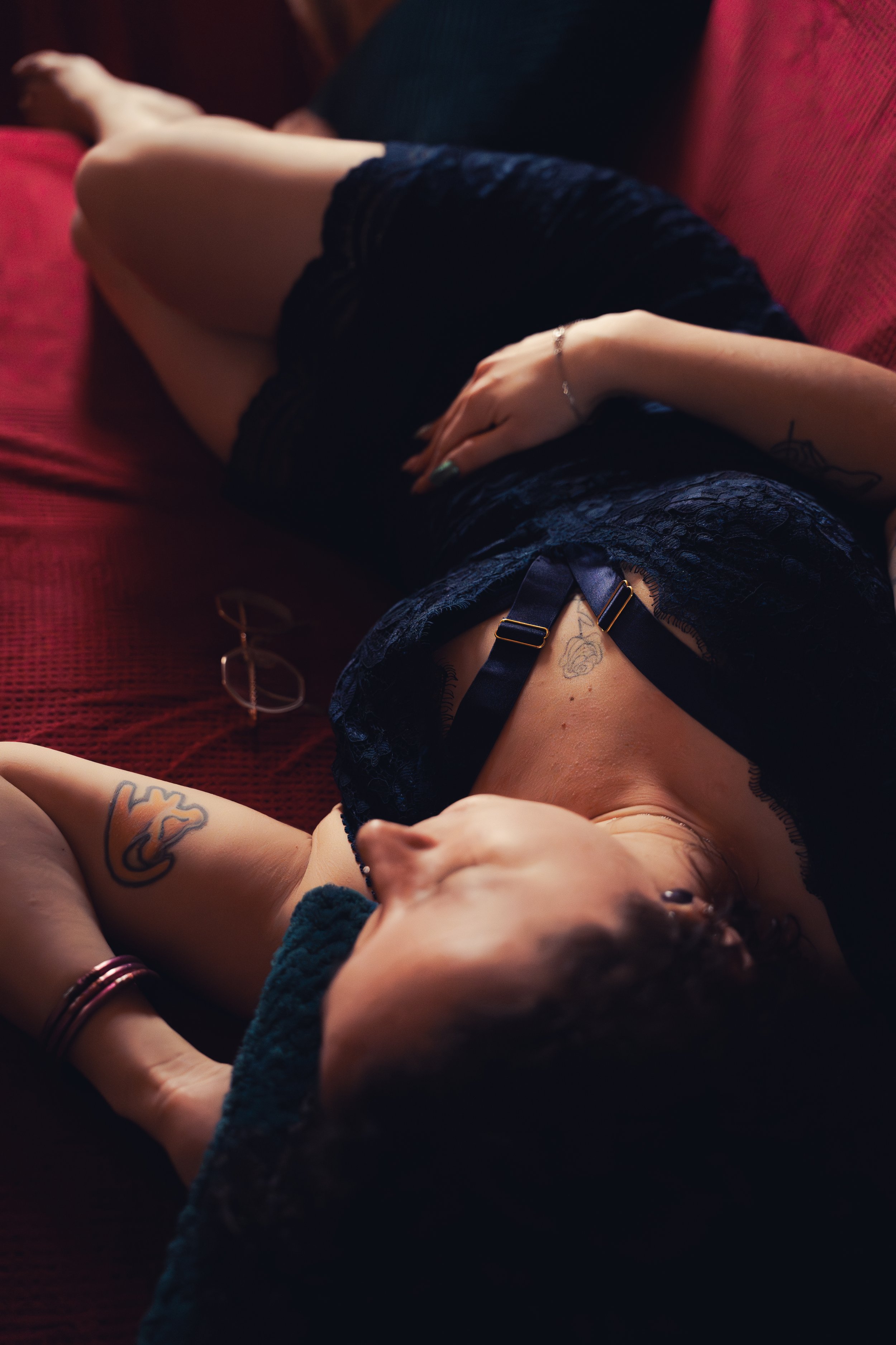 Femme allongée sur un canapé, portant une robe en dentelle noire et des tatouages visibles, avec una paire de lunettes à côté d'elle.