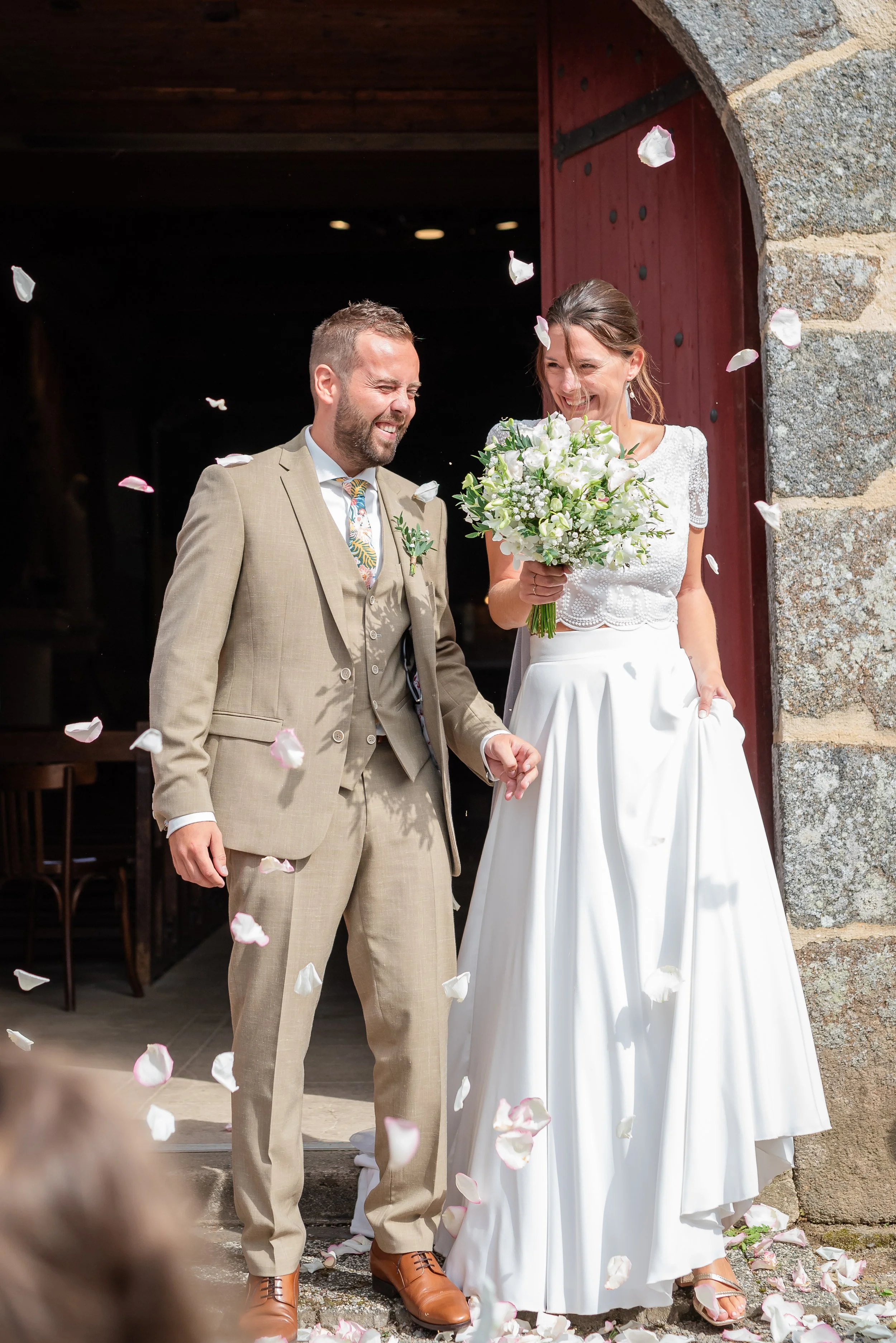 Un couple de mariés souriants sortant d'une église, entouré de pétales de fleurs, lors d'un mariage.