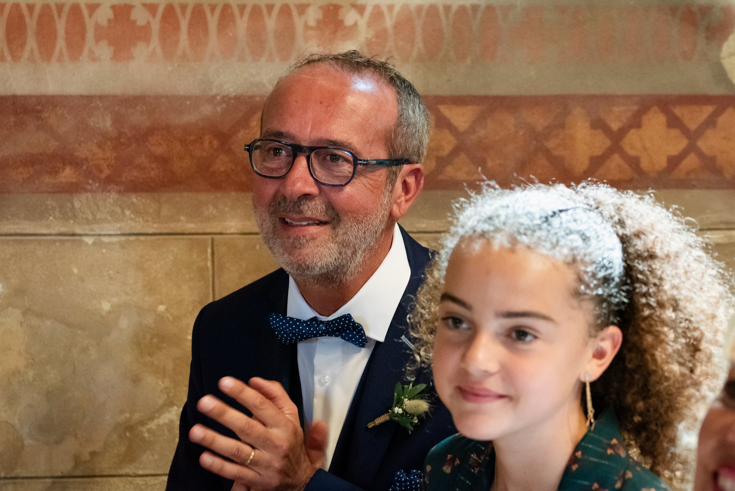 Homme âgé en costume noir avec nœud papillon bleu, portant des lunettes et un petit bouquet de fleurs, posant avec une enfant à cheveux bouclés et mousseux, dans un décor intérieur avec un mur en pierre