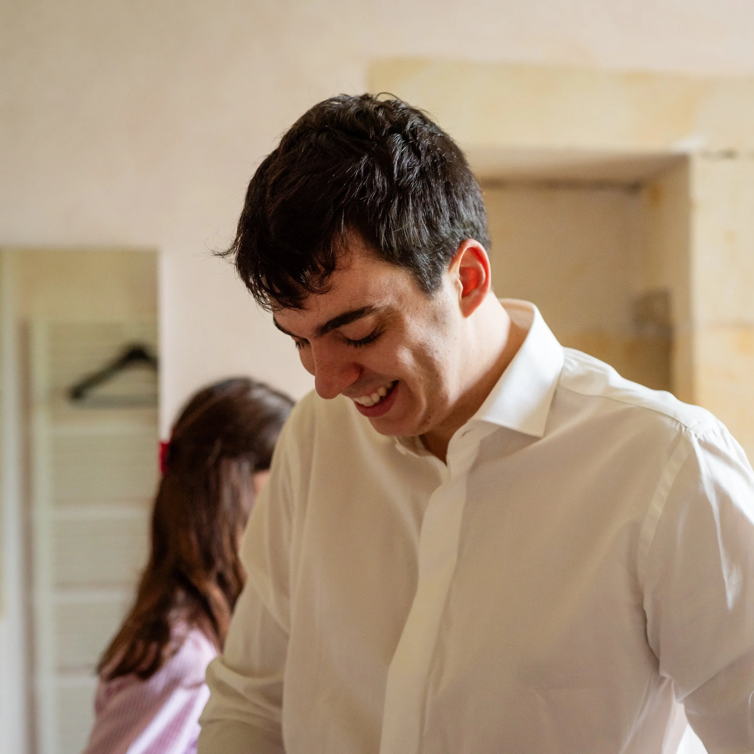 Un jeune homme souriant, avec des cheveux foncés et courts, portant une chemise blanche, regardant vers le bas dans un intérieur.