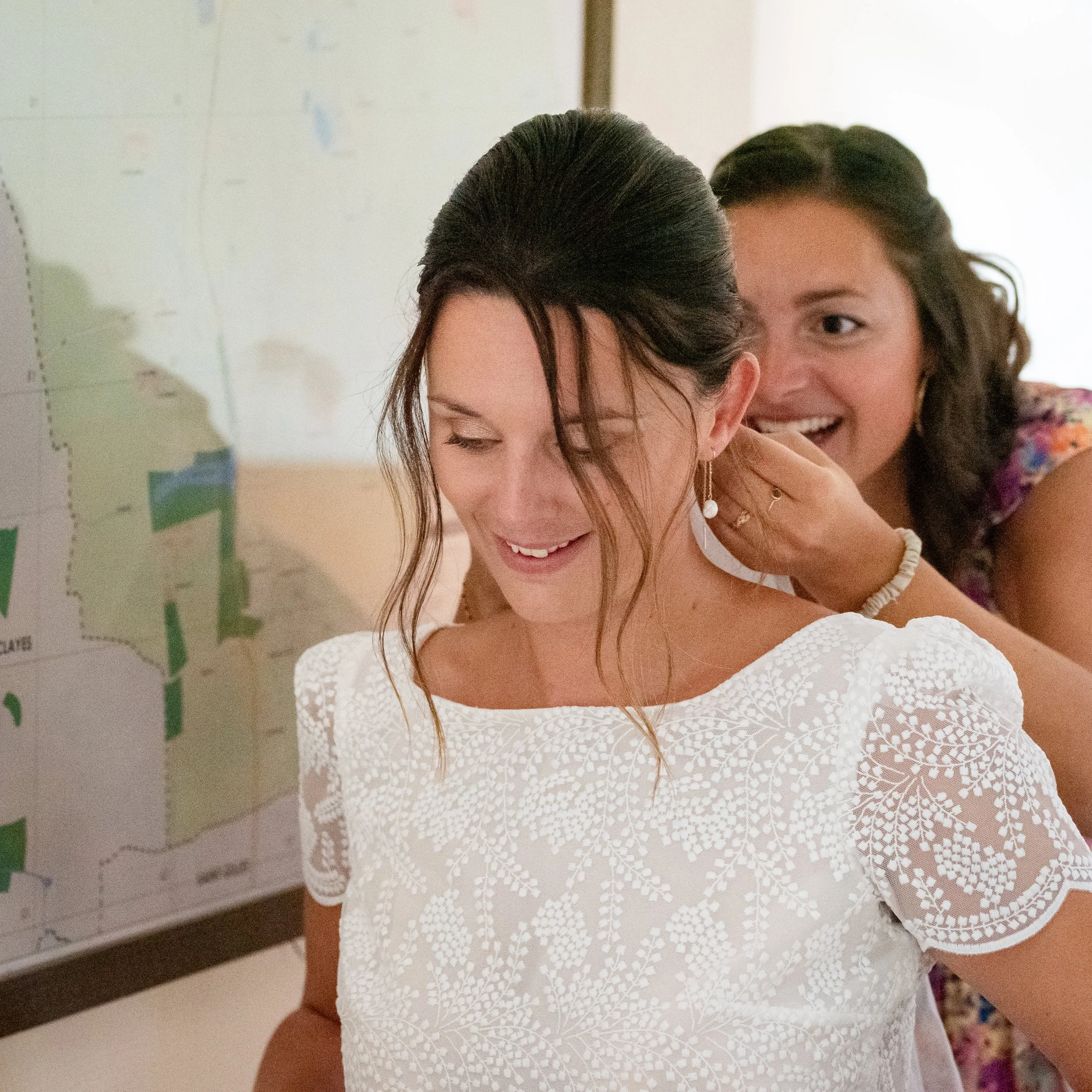 Deux jeunes femmes souriantes, l'une avec les yeux fermés et l'autre regardant, dans un environnement intérieur avec un tableau ou une carte au mur.