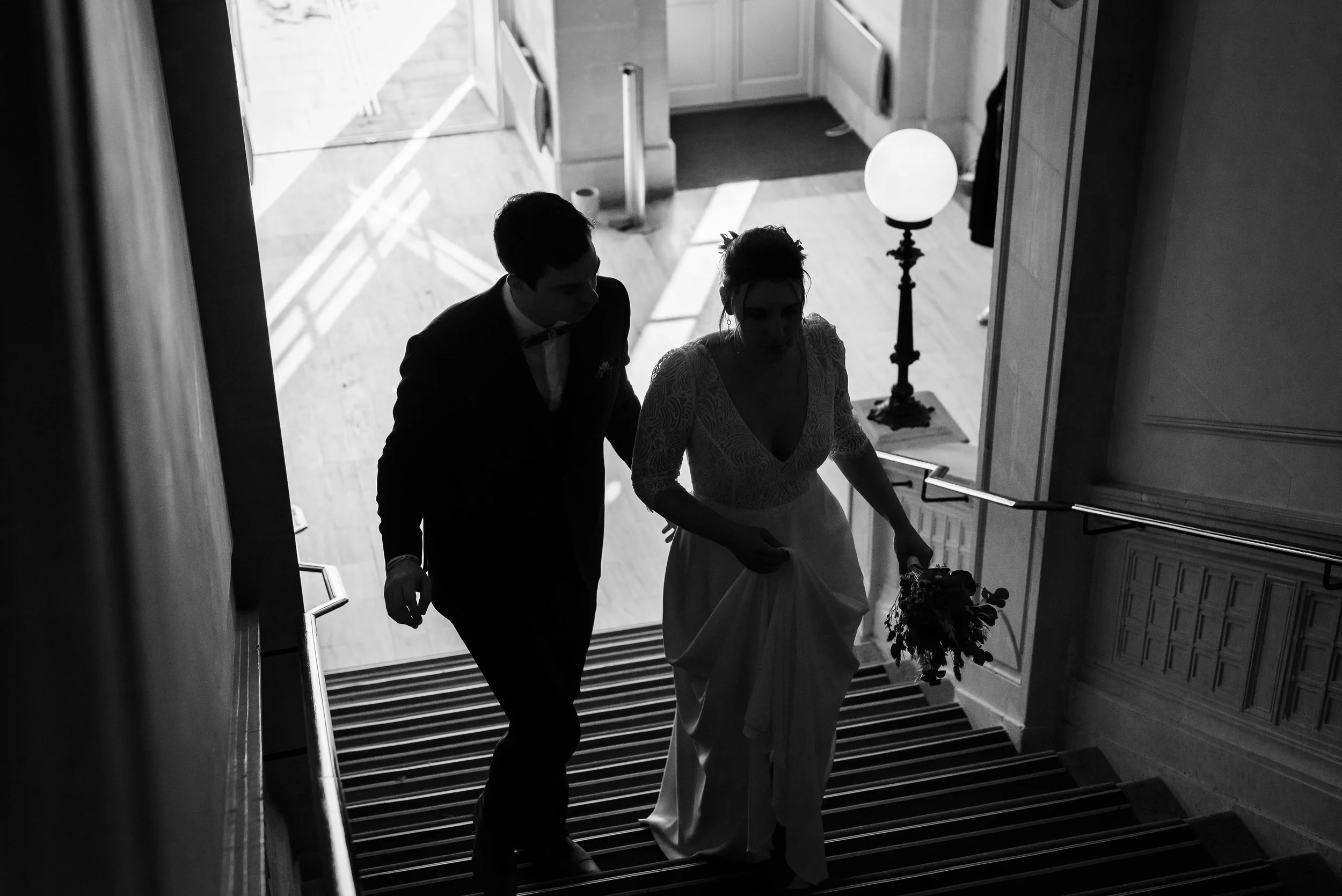 Deux personnes, un homme en costume et une femme en robe de mariée, montent un escalier en bois à l intérieur d’un bâtiment.
