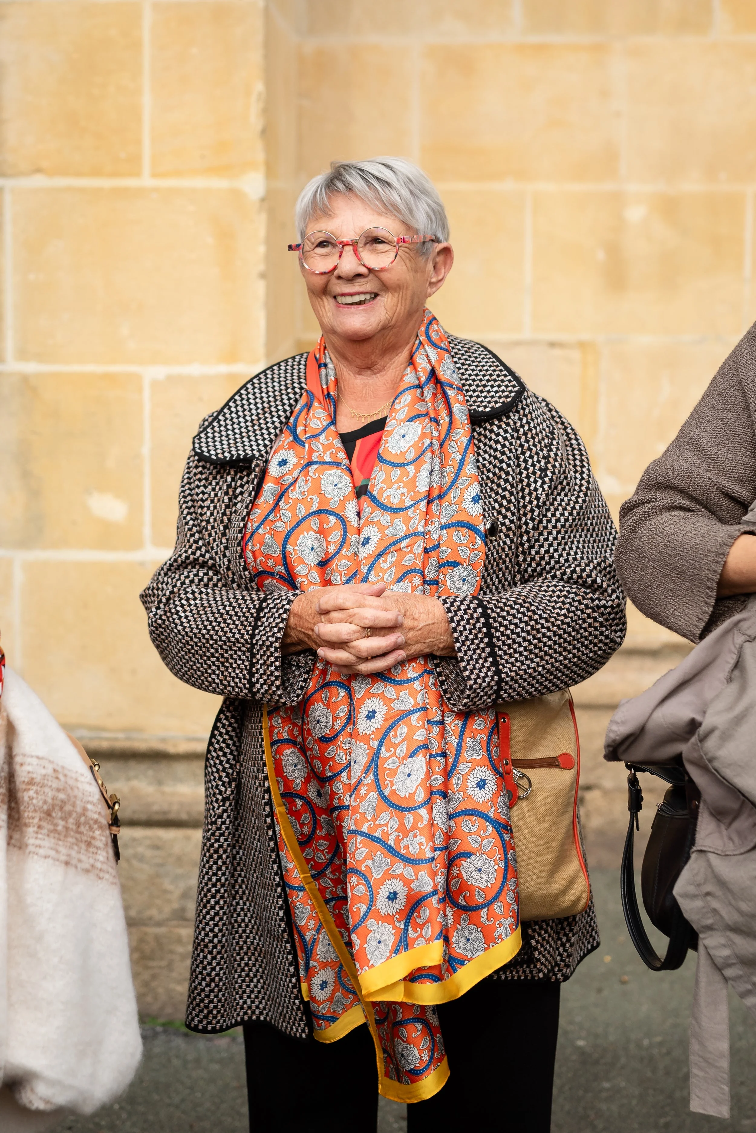 Une femme âgée souriante avec des cheveux gris courts, portant des lunettes à monture colorée, une veste à carreaux et une écharpe colorée avec un motif floral, avec un sac beige et un pantalon noir. Elle se tient devant un mur en pierre.