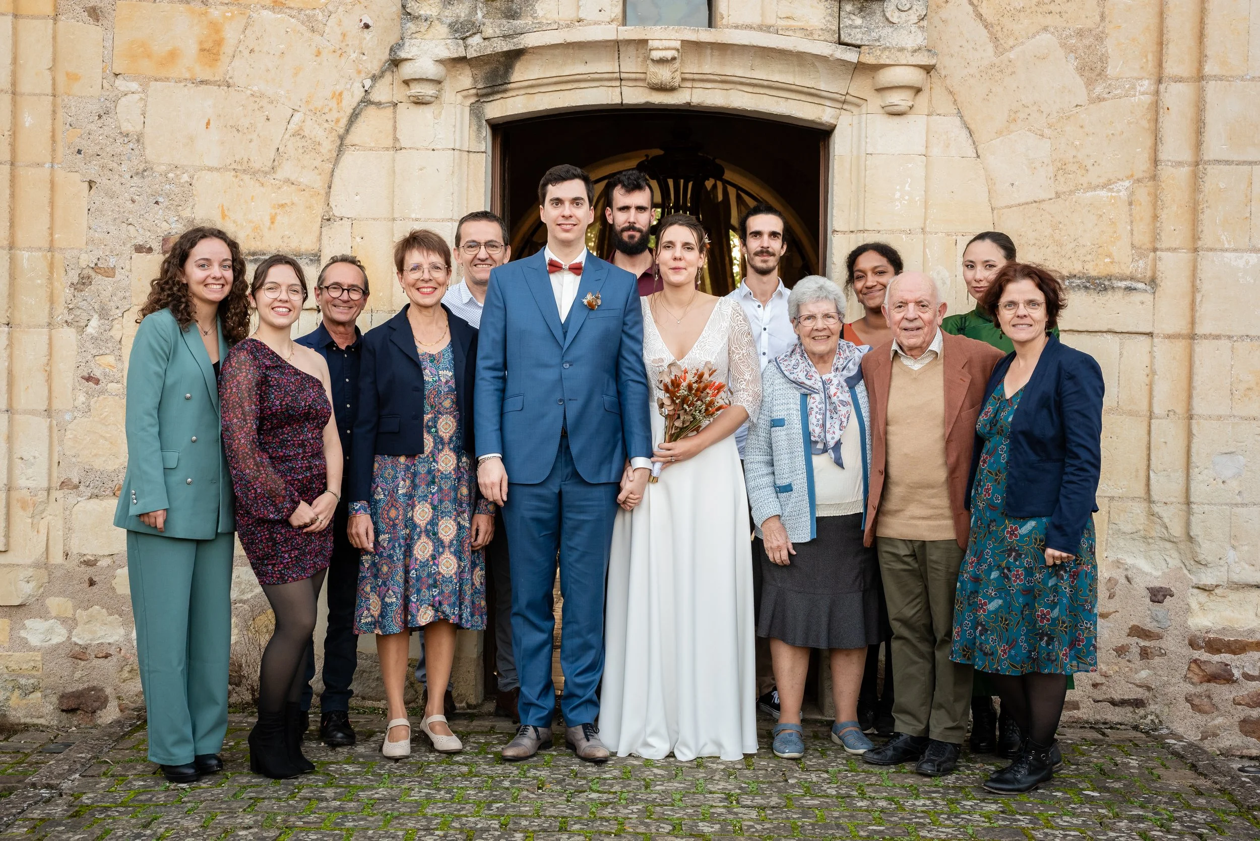 Groupe de personnes posant devant une ancienne bâtisse en pierre, comprenant un jeune homme en costume bleu, une femme en robe blanche, et plusieurs autres personnes d'âges variés, dans un style décontracté et élégant.