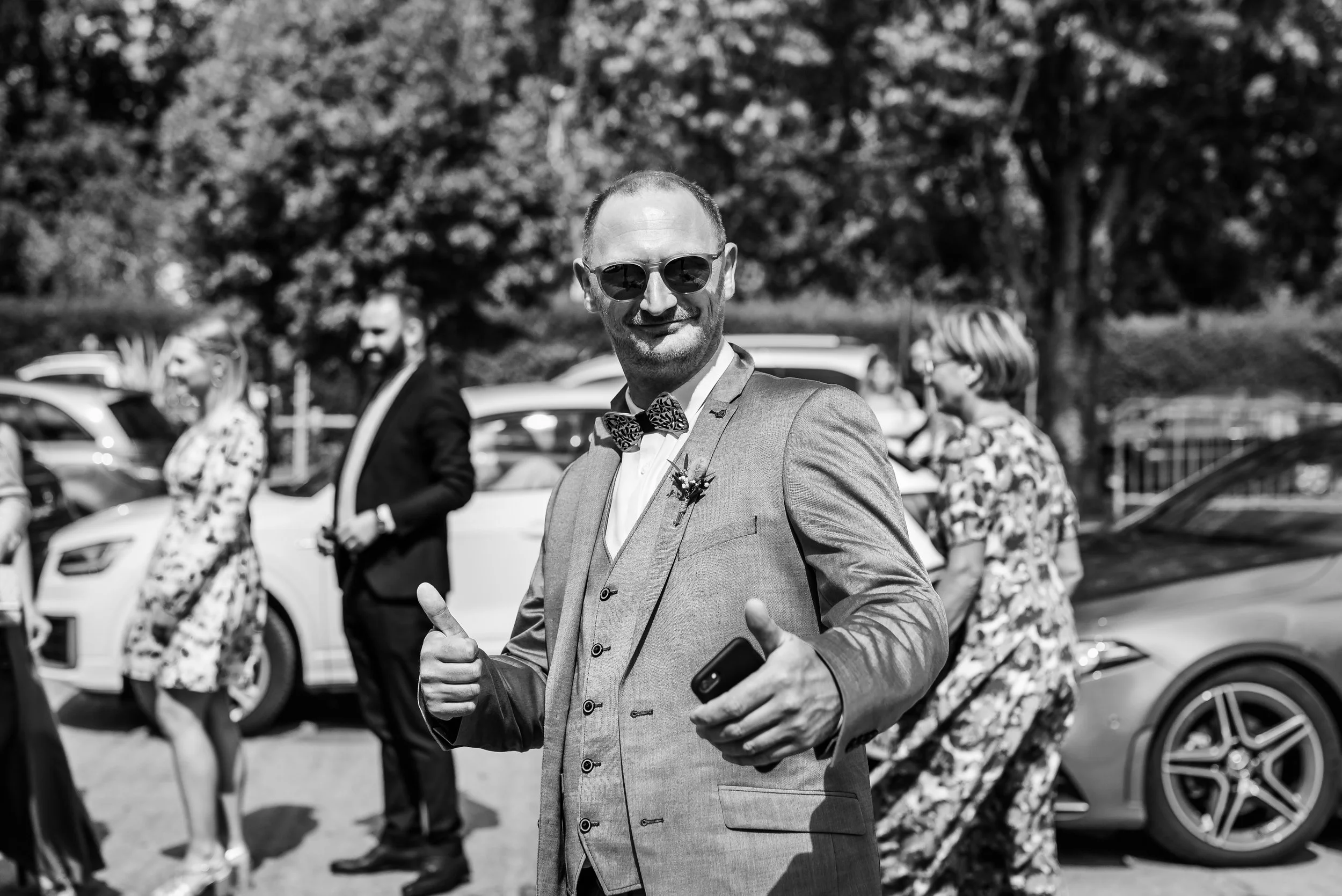 Un homme en costume avec des lunettes de soleil souriant et faisant un geste de pouce levé, lors d'un événement extérieur, avec plusieurs personnes et voitures en arrière-plan.