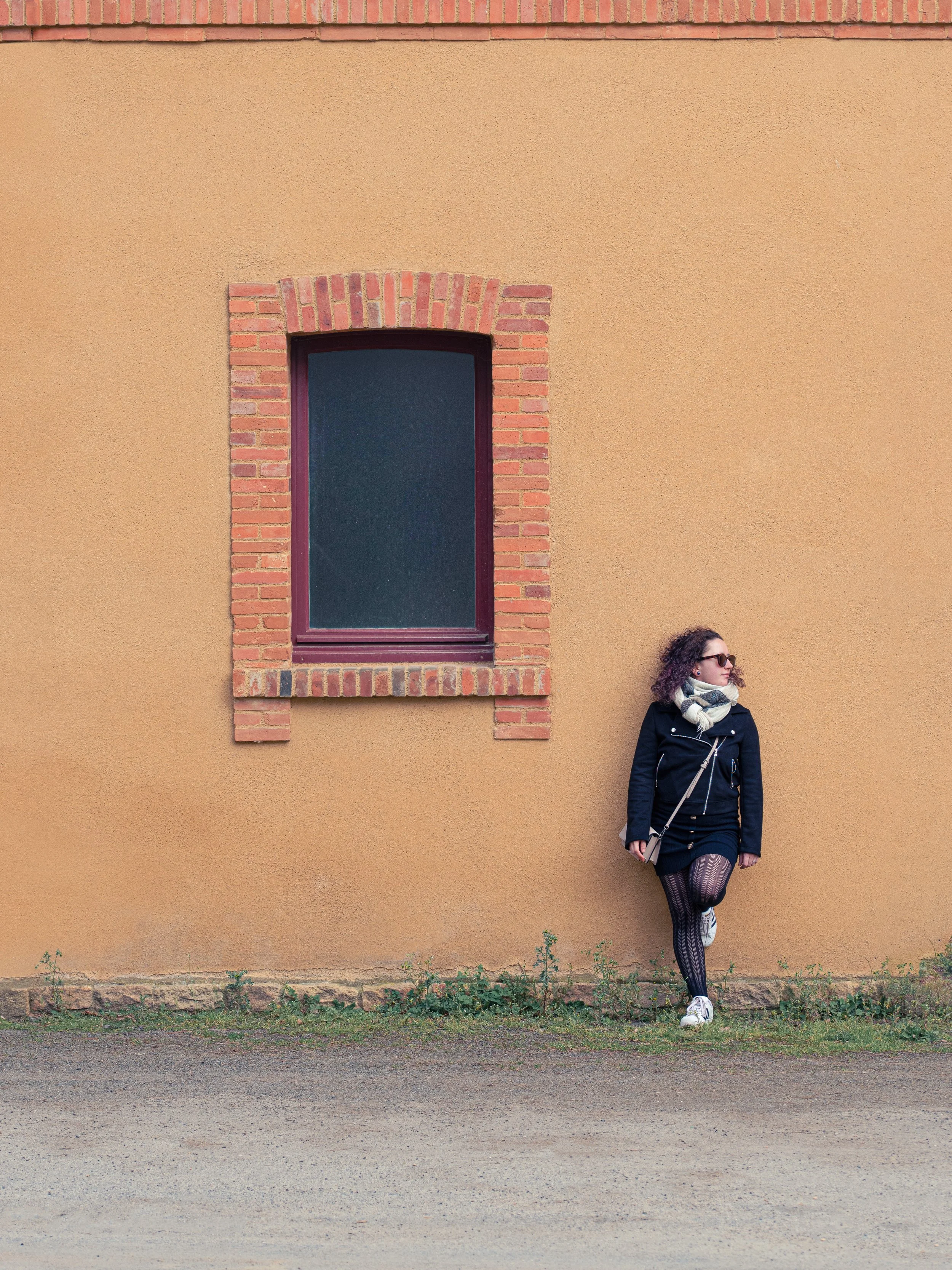 Une femme avec des cheveux bouclés portant des lunettes de soleil, un manteau noir, un foulard beige et une jupe à rayures, se tient contre un mur jaune avec une fenêtre encadrée en briques, en position décontractée avec une jambe croisée.