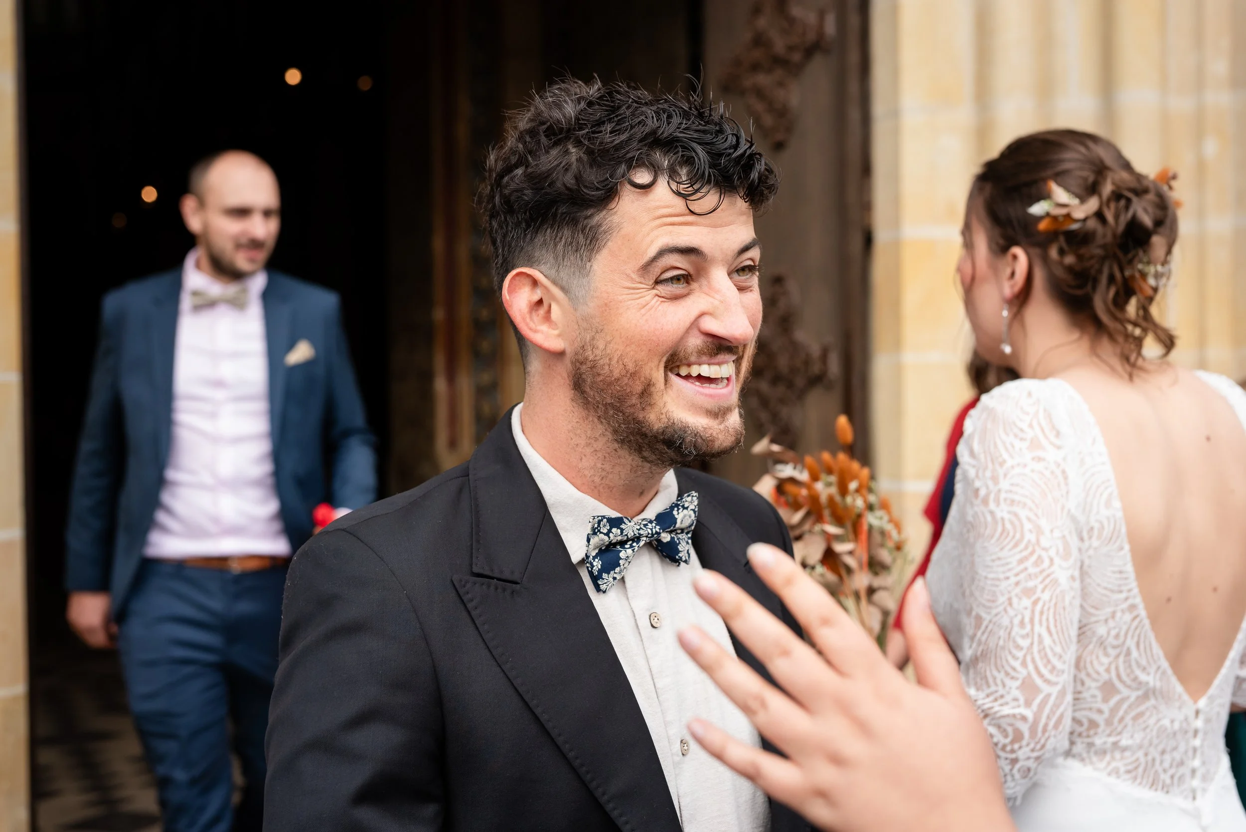 Un homme habillé en costume noir avec une chemise blanche et un nœud papillon à motif floral sourit lors d'une cérémonie de mariage, face à une femme dont le visage n'est pas visible.
