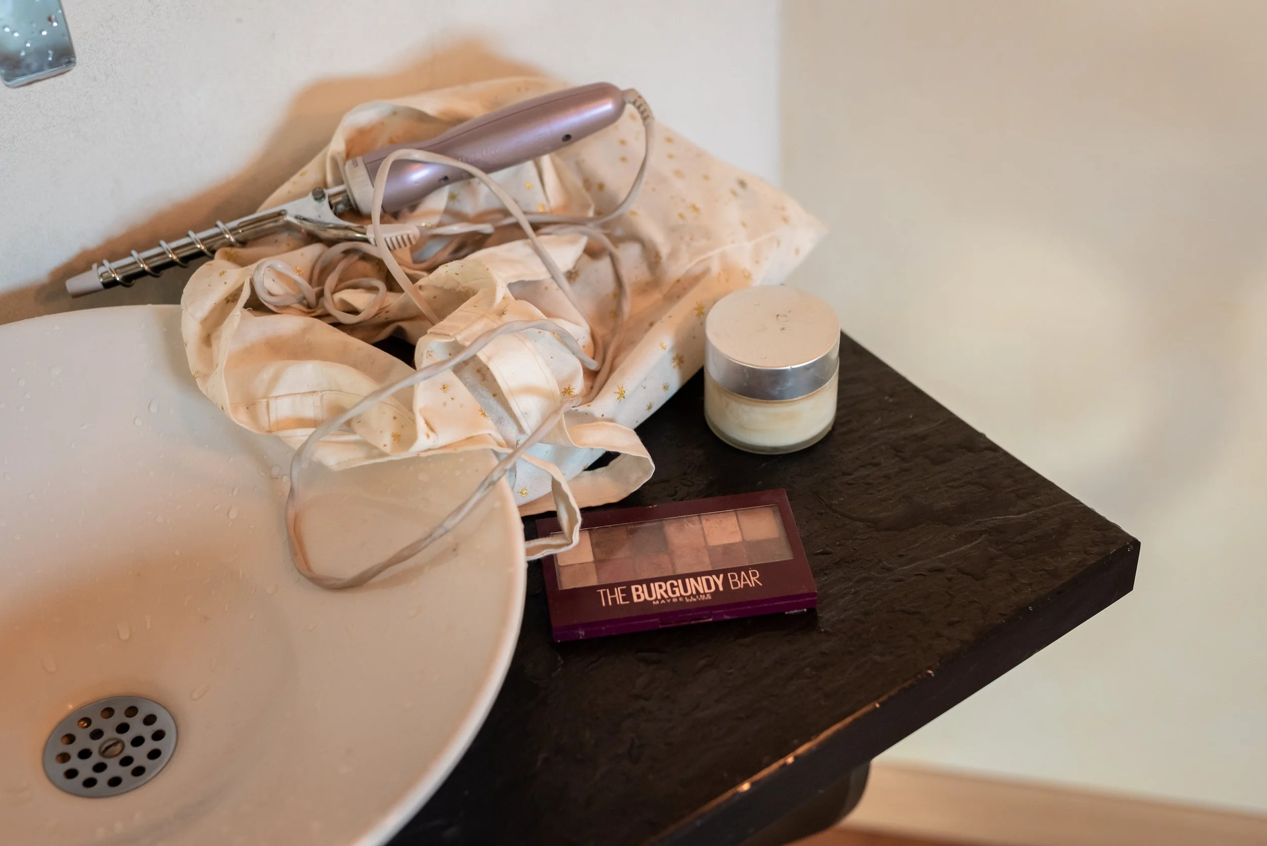 Une montre de beauté avec un dispositif électrique posé sur un tissu avec des motifs d'étoiles, à côté d'un pot de crème, posé sur une surface en bois. On voit aussi un petit maquillage harmonieux avec des nuances de marron et de rose.