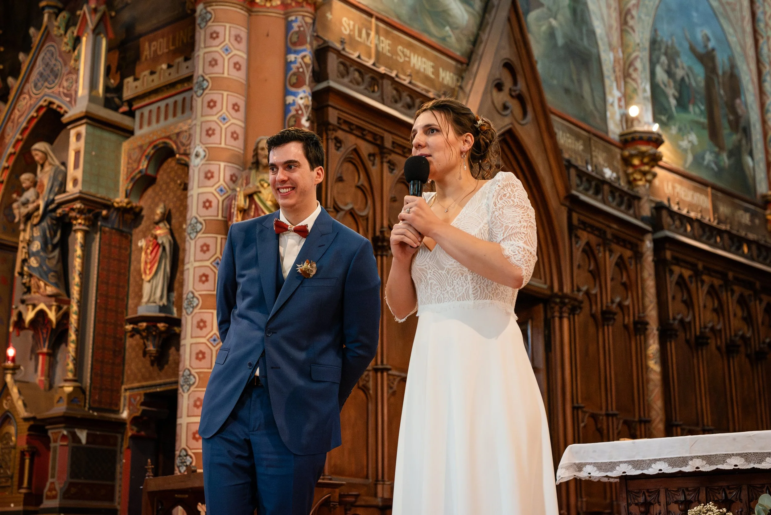 Un couple lors de leur mariage dans une église, la femme tient un micro et parle, l'homme porte un costume bleu avec un nœud papillon rouge, ils sont devant un décor d'autel richement orné.