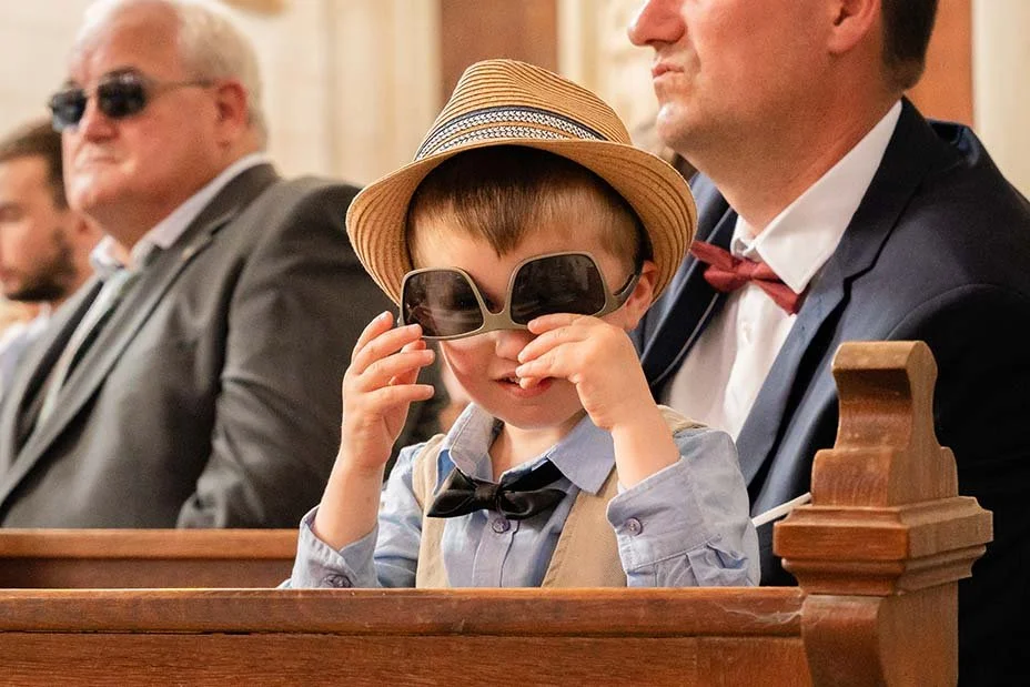 Un jeune garçon portant un chapeau, des lunettes de soleil sur le nez, une chemise avec un nœud papillon, assis dans une église ou une salle de cérémonie, entouré d'adultes.