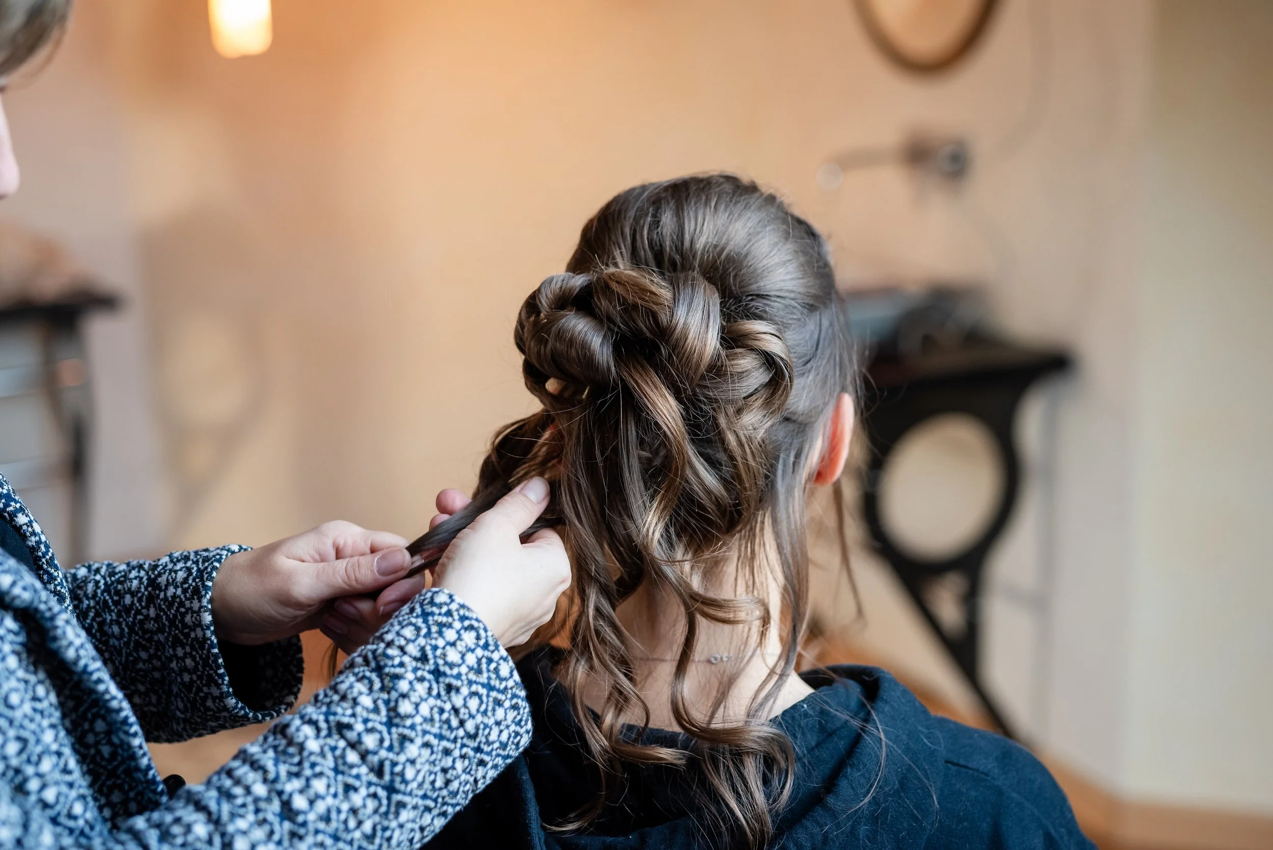 Une femme coiffe une autre personne, qui a de longs cheveux bruns. La scène se déroule en intérieur, avec un mur beige et quelques meubles en arrière-plan.