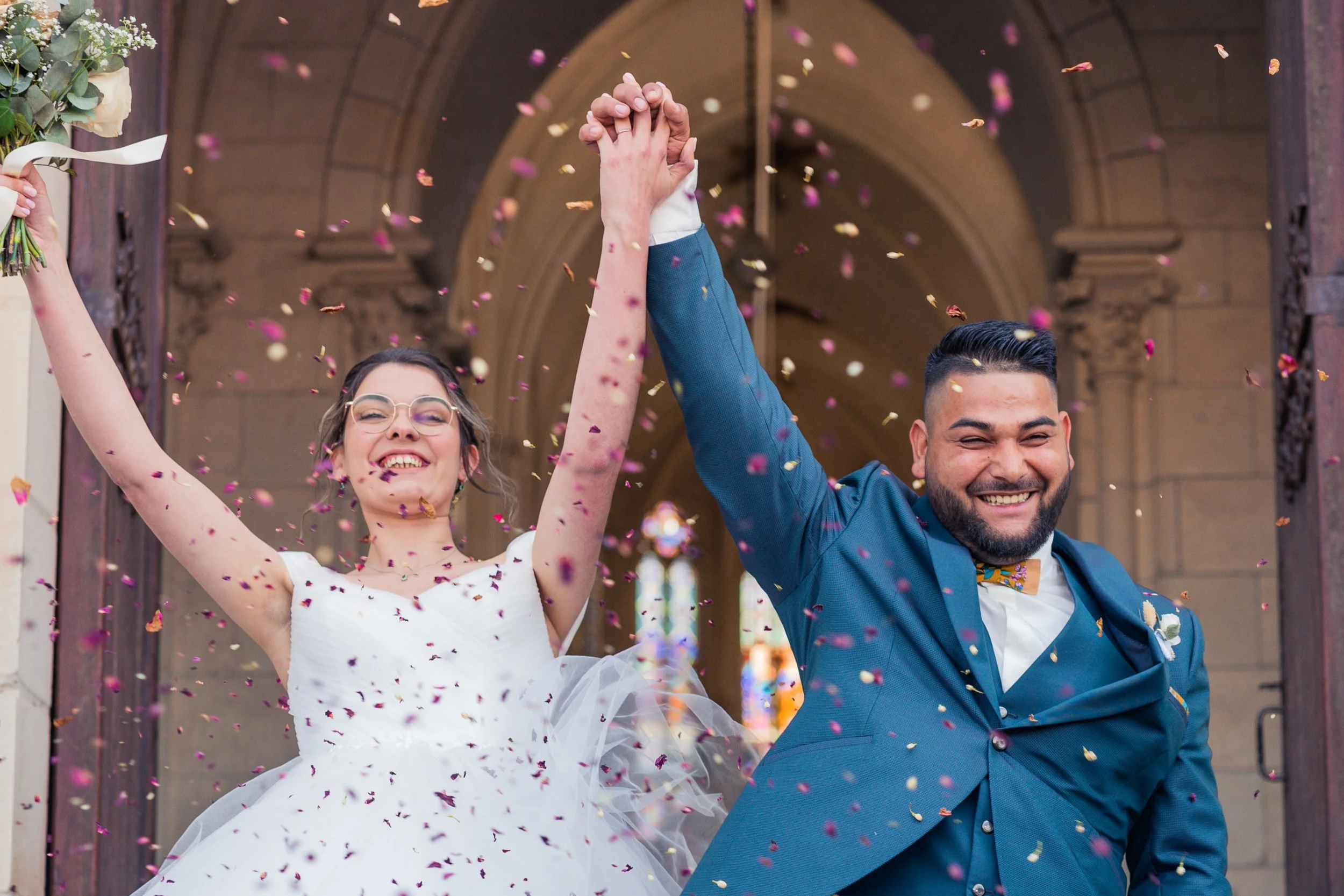 Un couple de mariés célèbre leur mariage avec des confettis, ils se tiennent par la main et sourient devant une église en pierre, avec des vitraux colorés en arrière-plan.