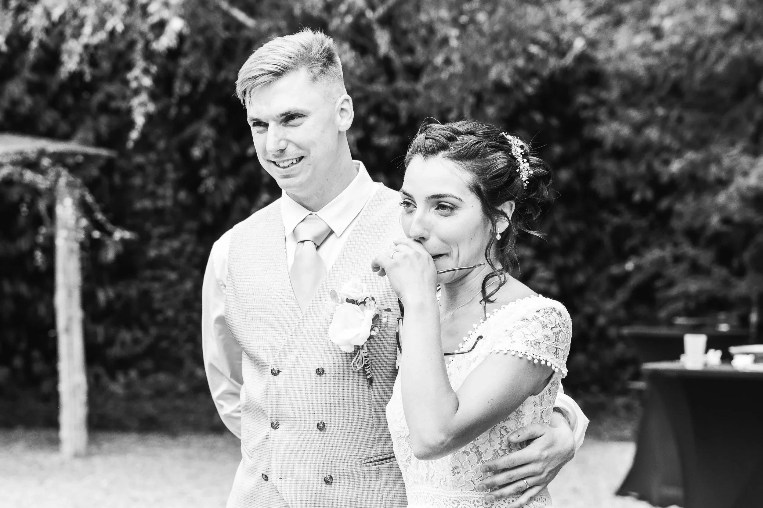 Un couple lors d'une cérémonie de mariage en plein air, l'homme en costume clair et la femme en robe de mariée avec un voile, partage un moment émouvant et joyeux.