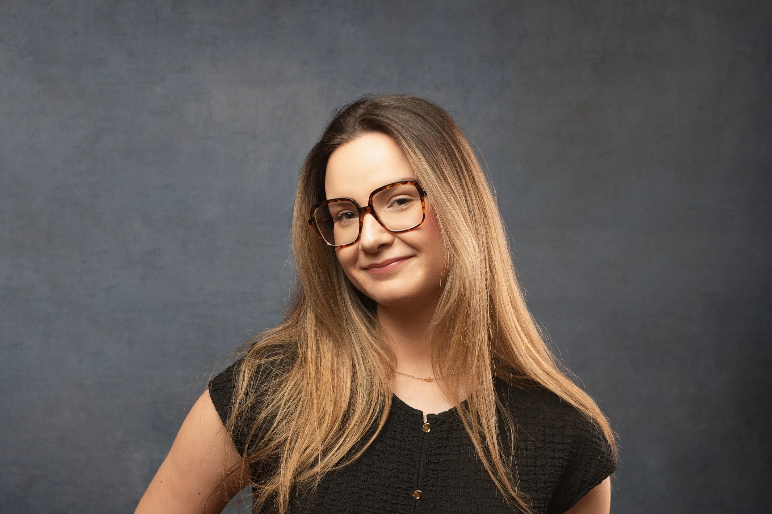Jeune femme souriante portant des lunettes à gros cadre tacheté de tortue, avec des cheveux longs et marron clair, vêtue d'un haut noir texturé, posant devant un fond gris.