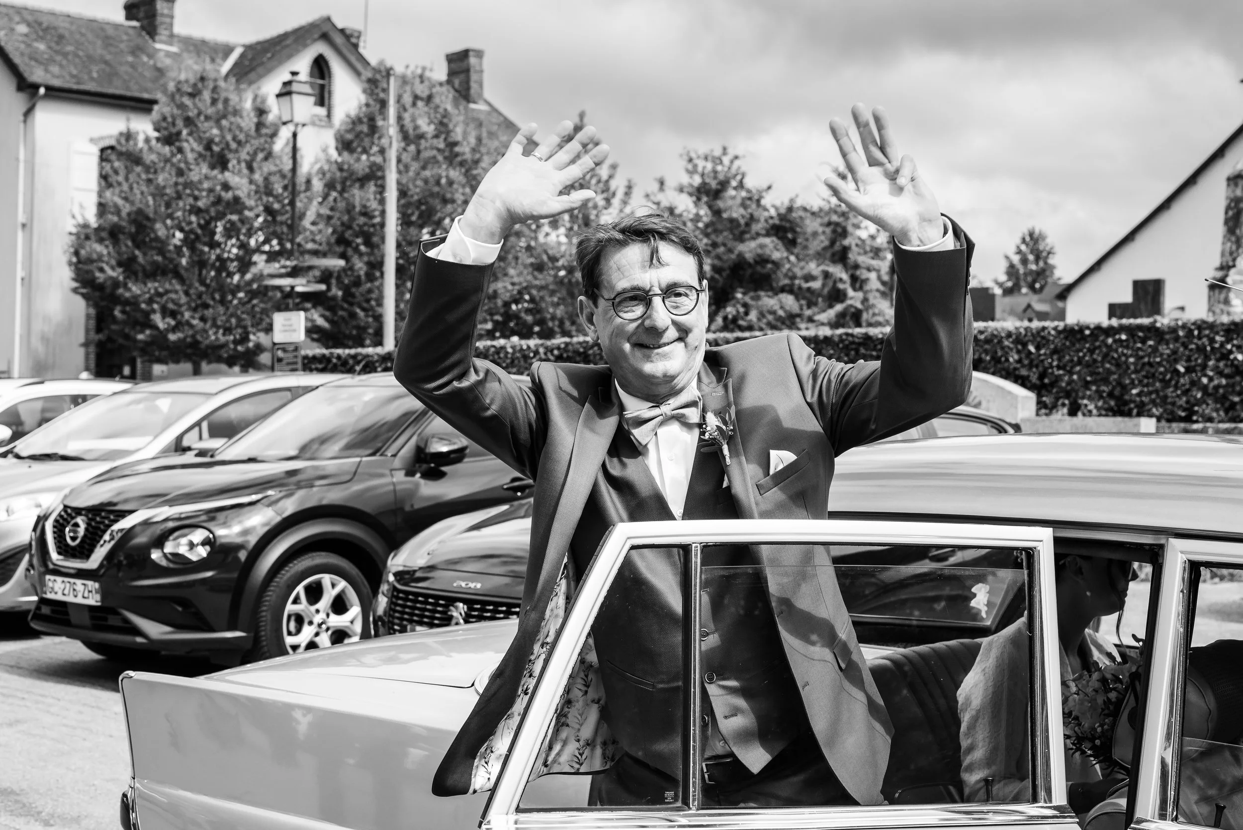 Homme souriant en costume et lunettes, saluant avec les deux mains, debout à côté d'une voiture décapotable aux portes ouvertes, dans un parking en pleine journée.
