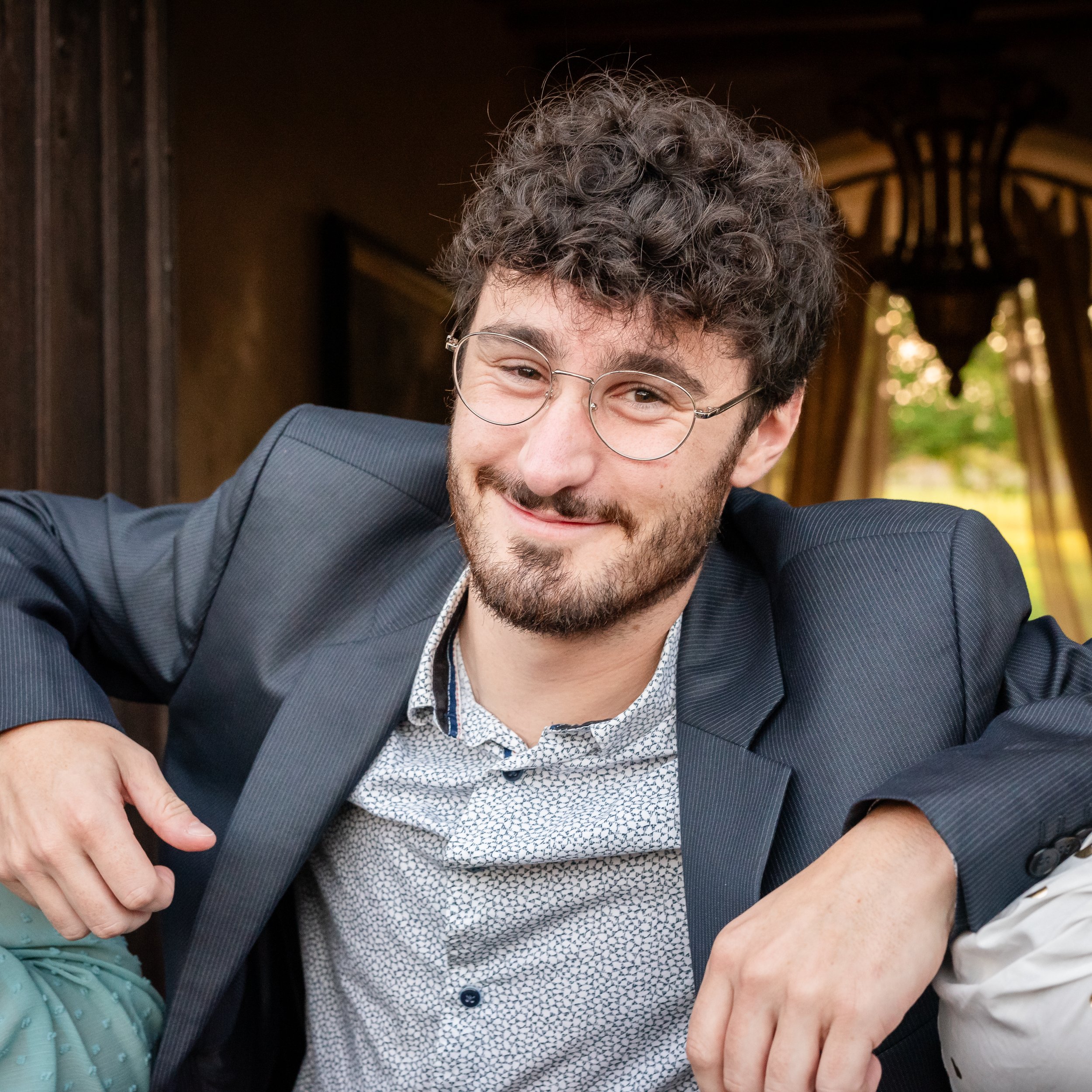 Un homme souriant portant des lunettes, habillé d'une veste bleue avec des motifs, dans un cadre intérieur avec un arrière-plan en bois et une lumière naturelle passant par une fenêtre.