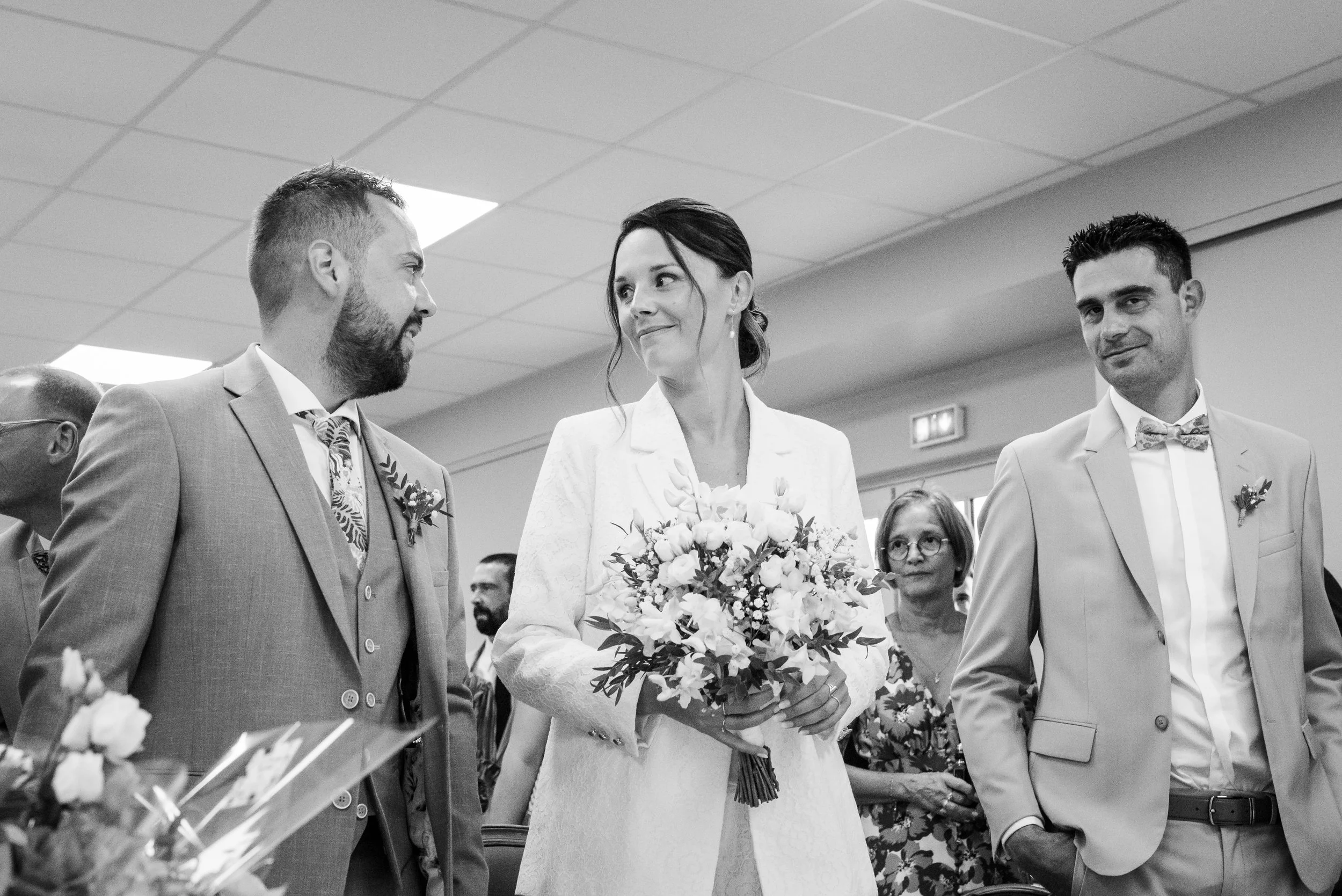 Un couple de mariés lors de leur cérémonie, entourés d'amis et famille, avec la mariée tenant un bouquet de fleurs.
