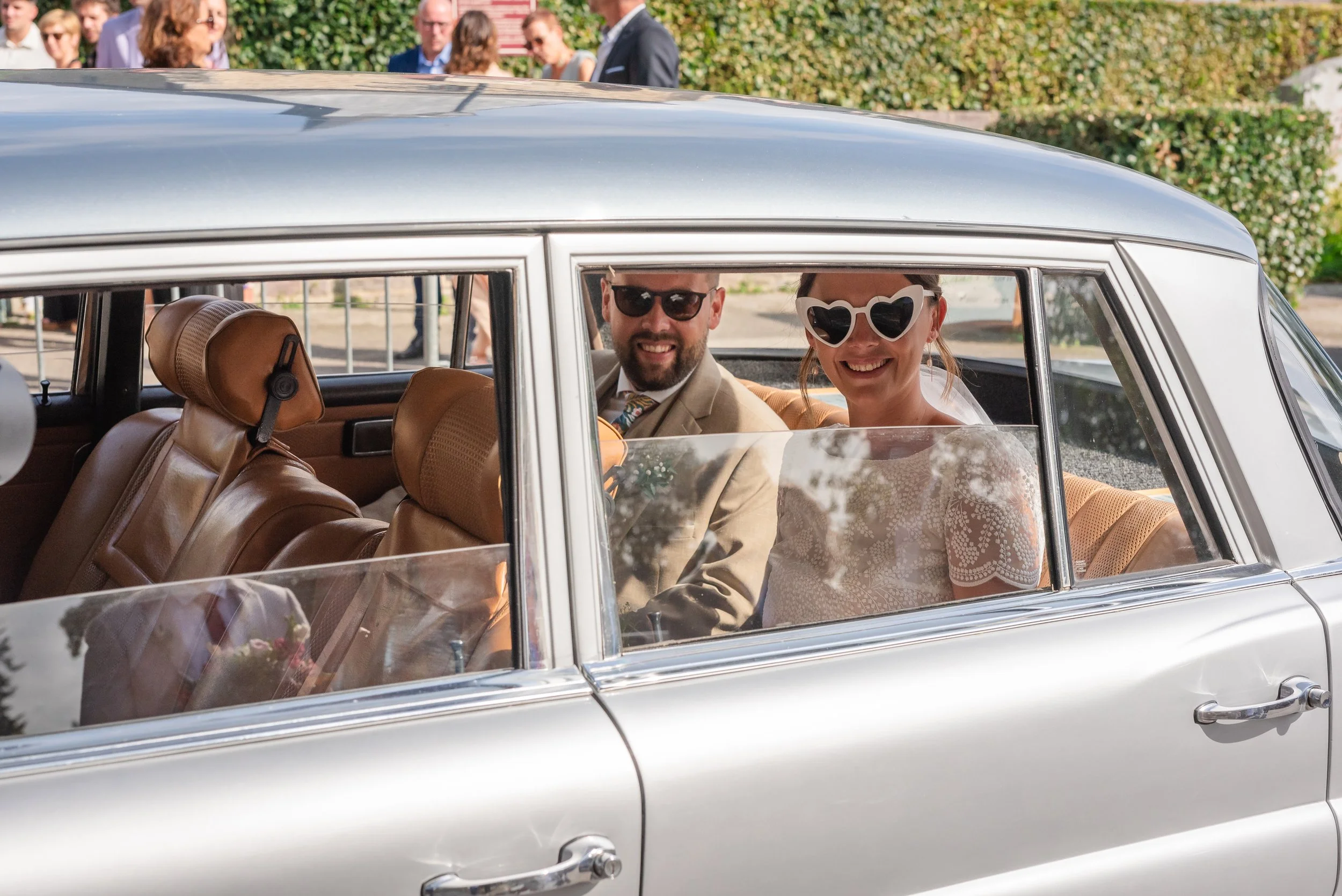 Un couple souriant dans une voiture classique, portant des lunettes de soleil, lors d'un événement en plein air avec un groupe de personnes en arrière-plan.