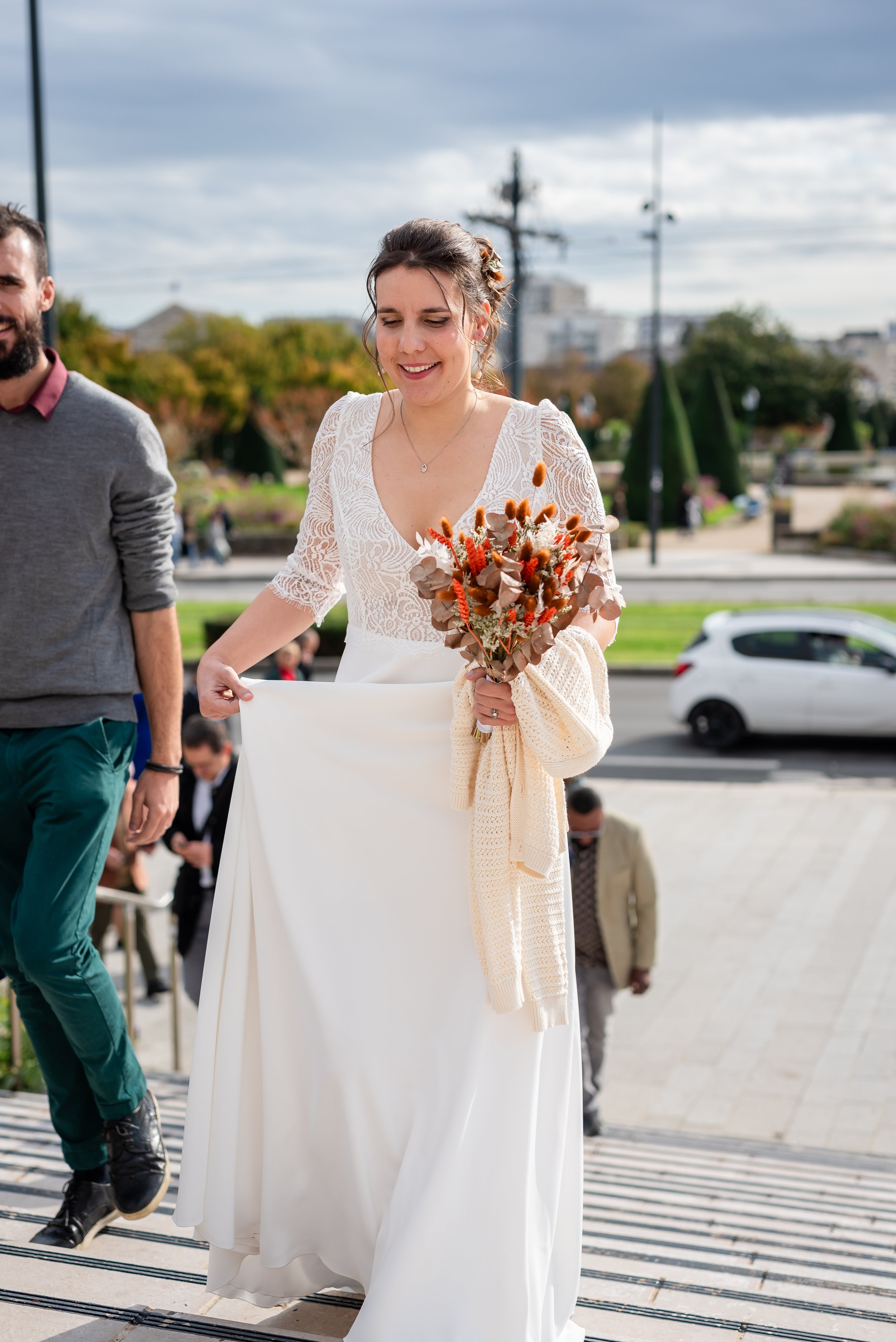 Une femme en robe blanche élégante avec un bouquet de fleurs orangées et brunes, marchant dans un espace public extérieur, avec plusieurs personnes en arrière-plan.