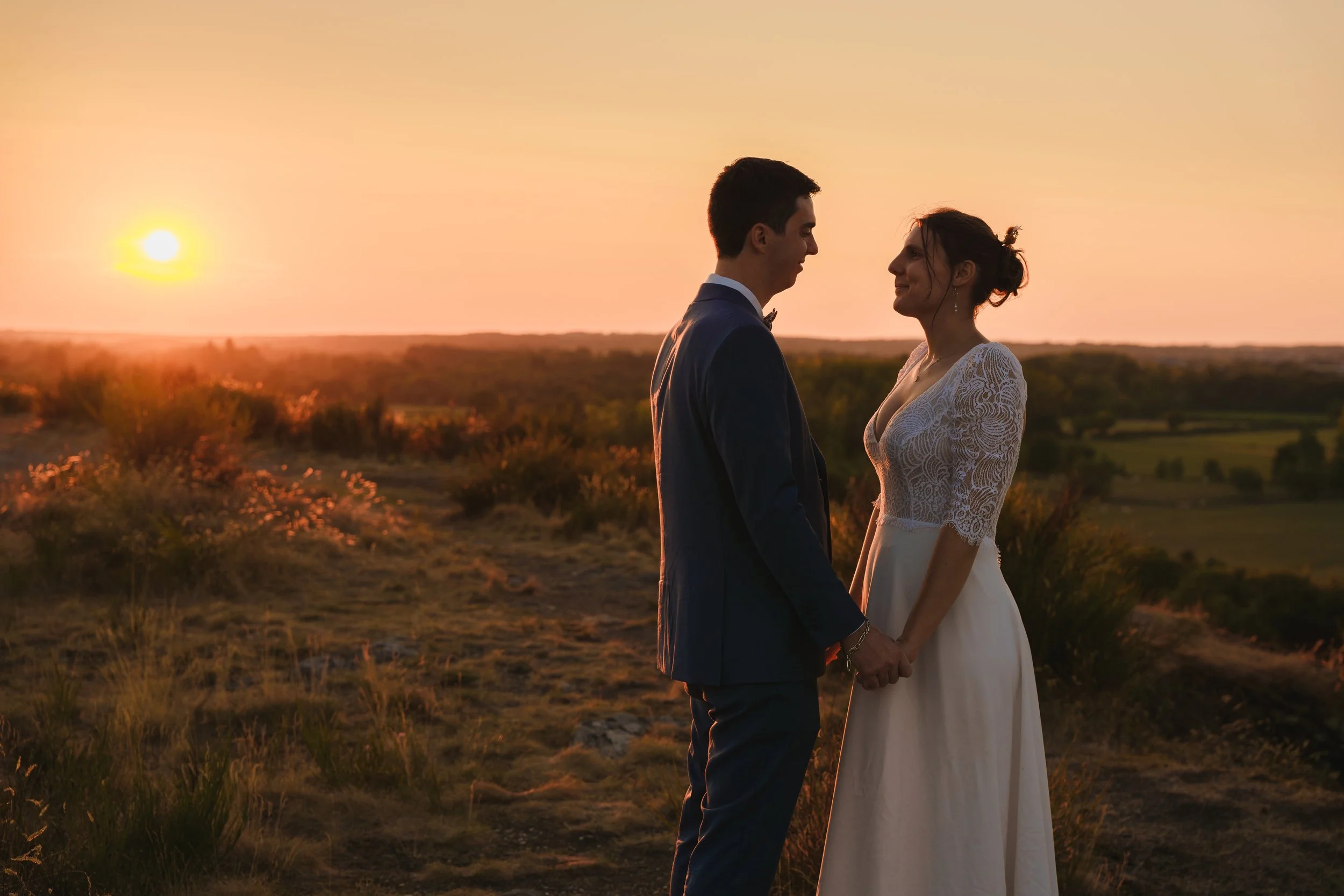 Un couple marié, un homme en costume et une femme en robe blanche, se tenant la main lors d'une cérémonie de mariage en plein air au coucher du soleil, avec un paysage rural en arrière-plan.