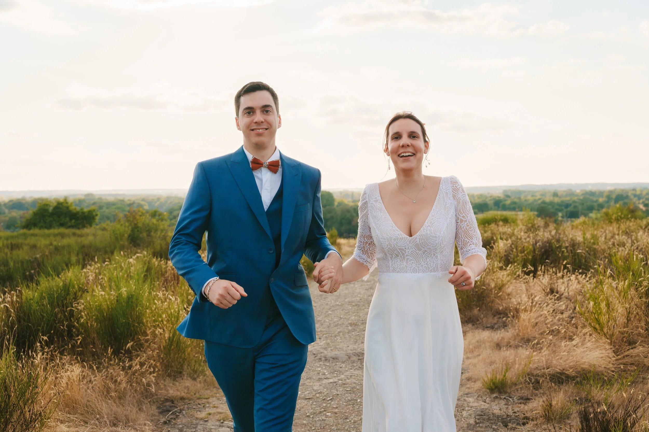 Un couple marchant main dans la main dans un paysage rural au coucher du soleil, la femme en robe blanche et l'homme en costume bleu avec un nœud papillon rouge.