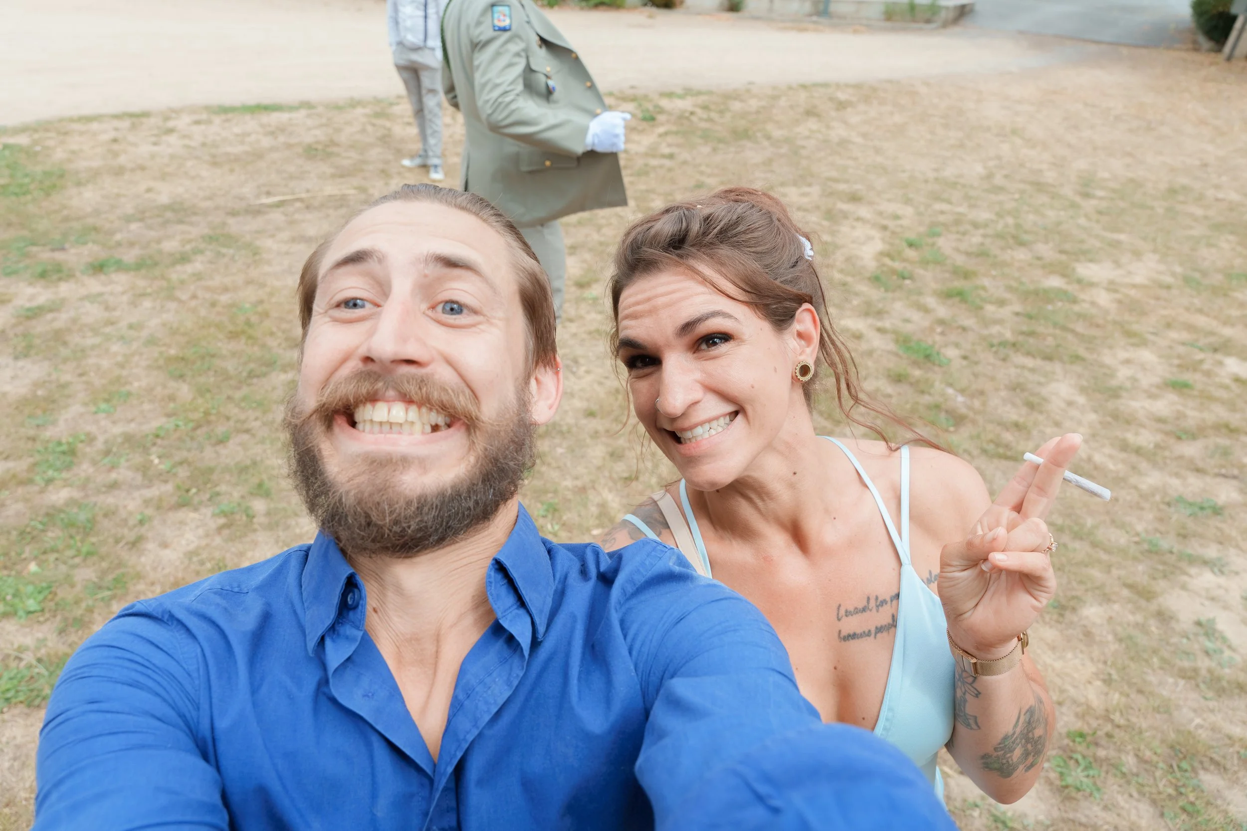 Deux adultes prennent un selfie dans un parc, l'homme a une barbe et porte une chemise bleue, la femme a des tatouages et tient une cigarette, ils sourient tous les deux.