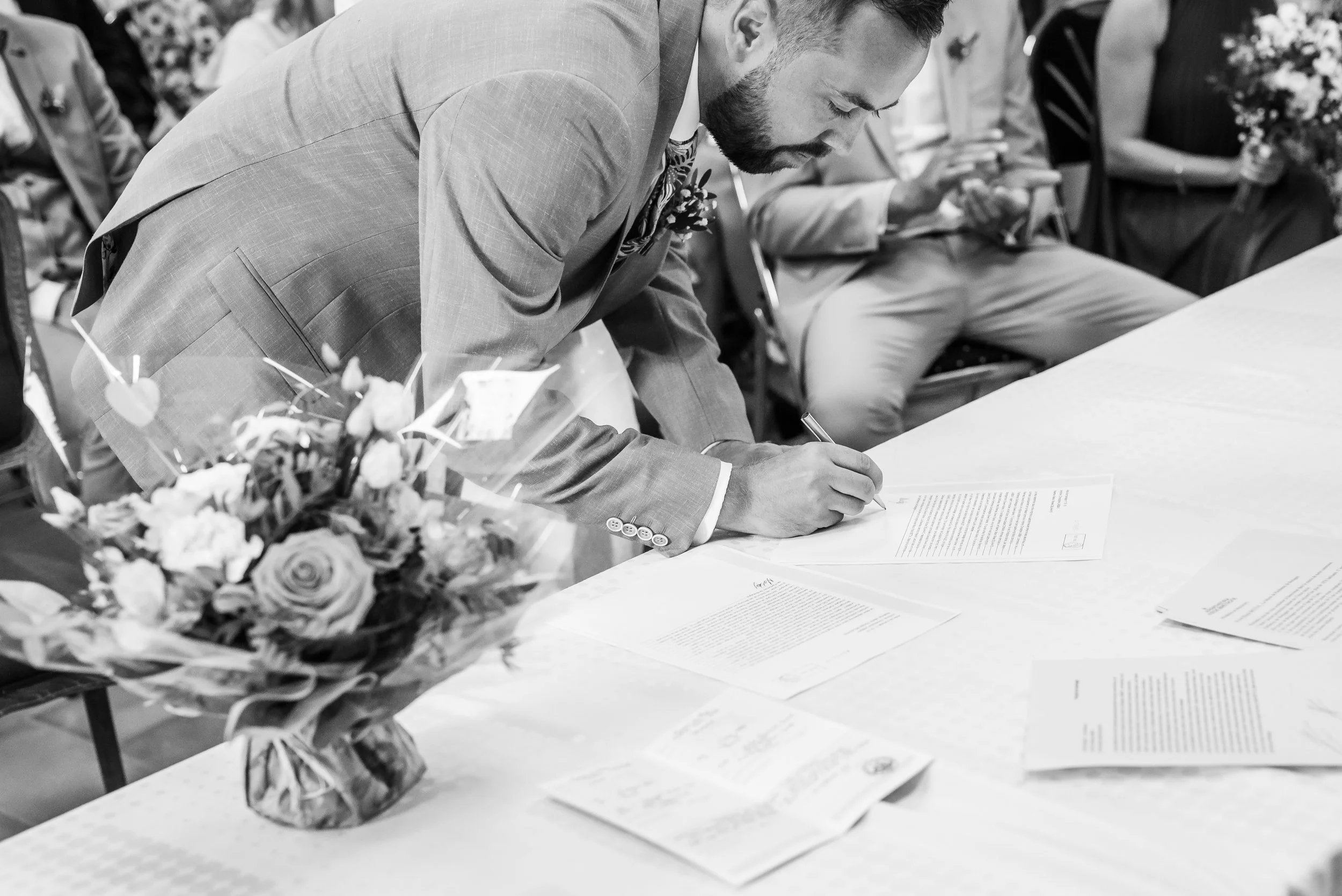 Un homme à la barbe signe un document à une table lors d'un événement, avec une bouquet de fleurs en premier plan.