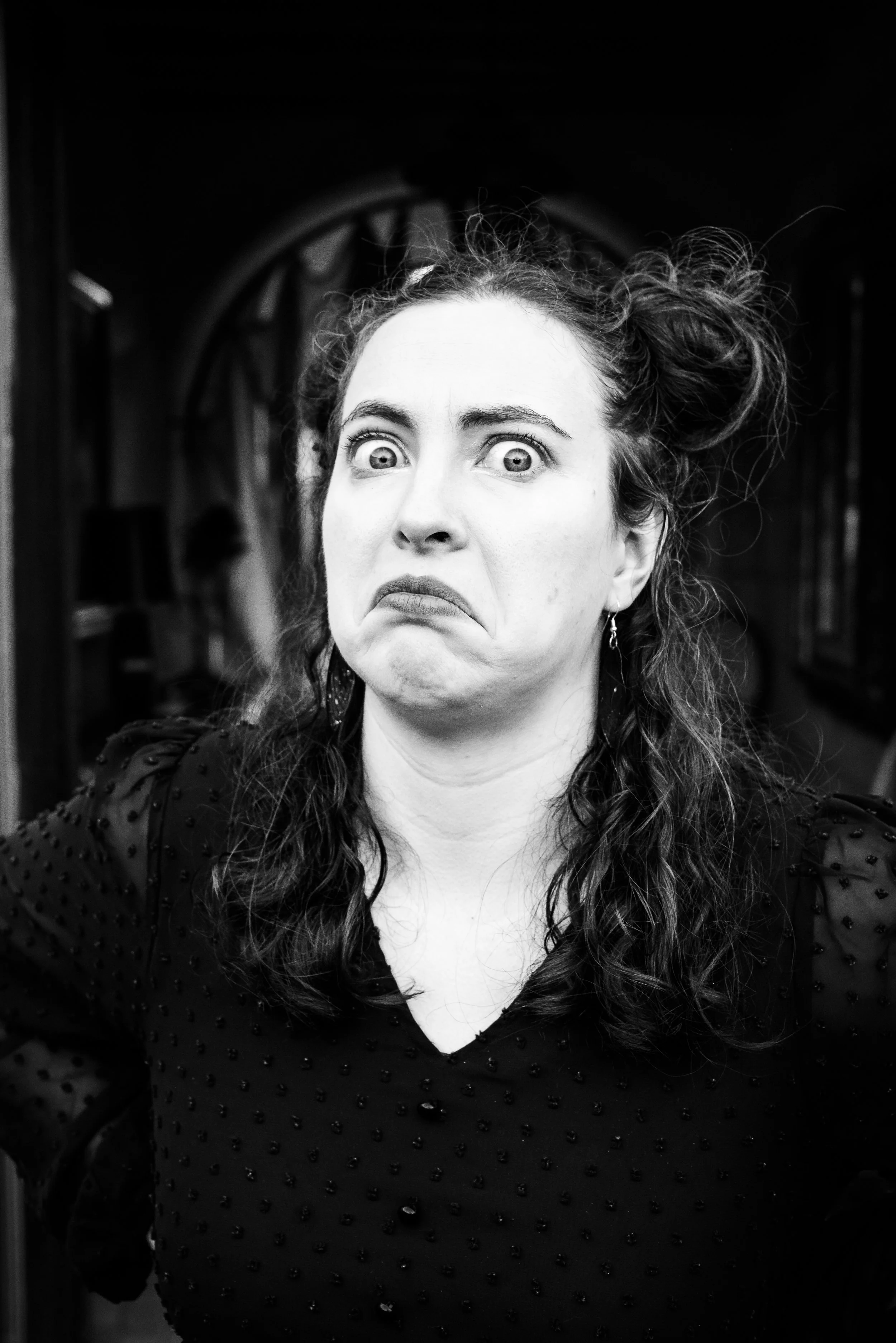 Femme avec expressions faciales exagérées, cheveux en désordre, portant une robe noire à pois, dans un décor sombre.