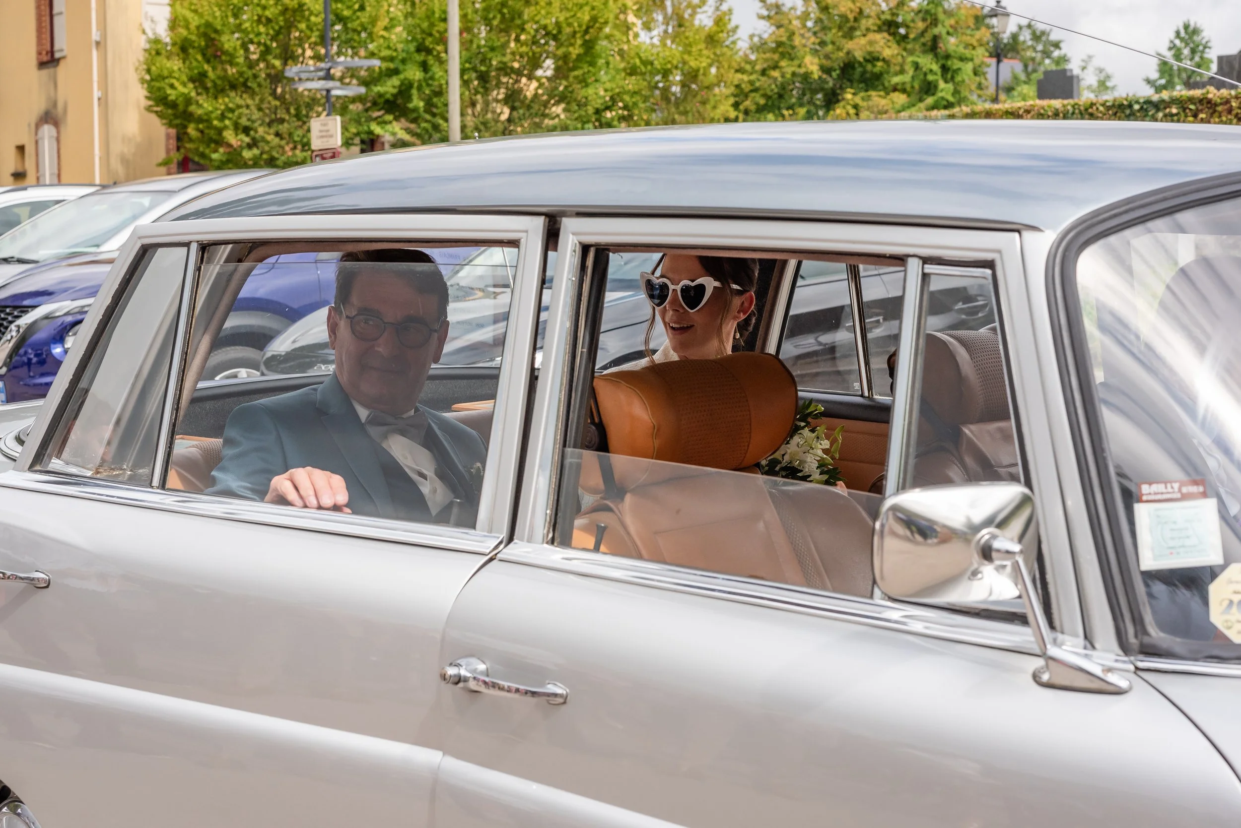 Un couple en costume assis dans une voiture vintage en pleine rue, avec la femme portant des lunettes de soleil en forme de cœur, lors d'une journée ensoleillée.