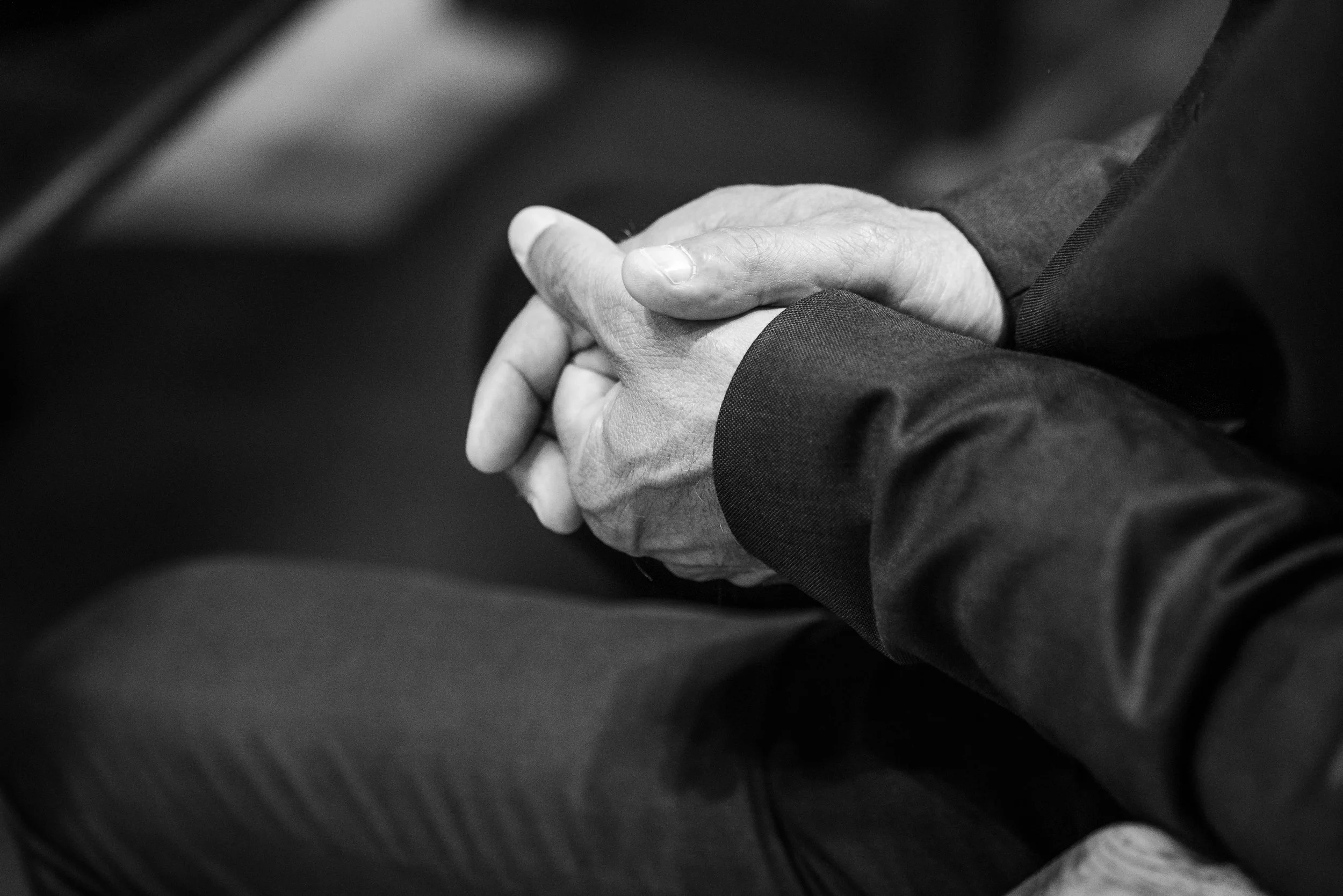 Mains croisées en position de prière ou de réflexion, vêtues d'un costume sombre, photographie en noir et blanc.
