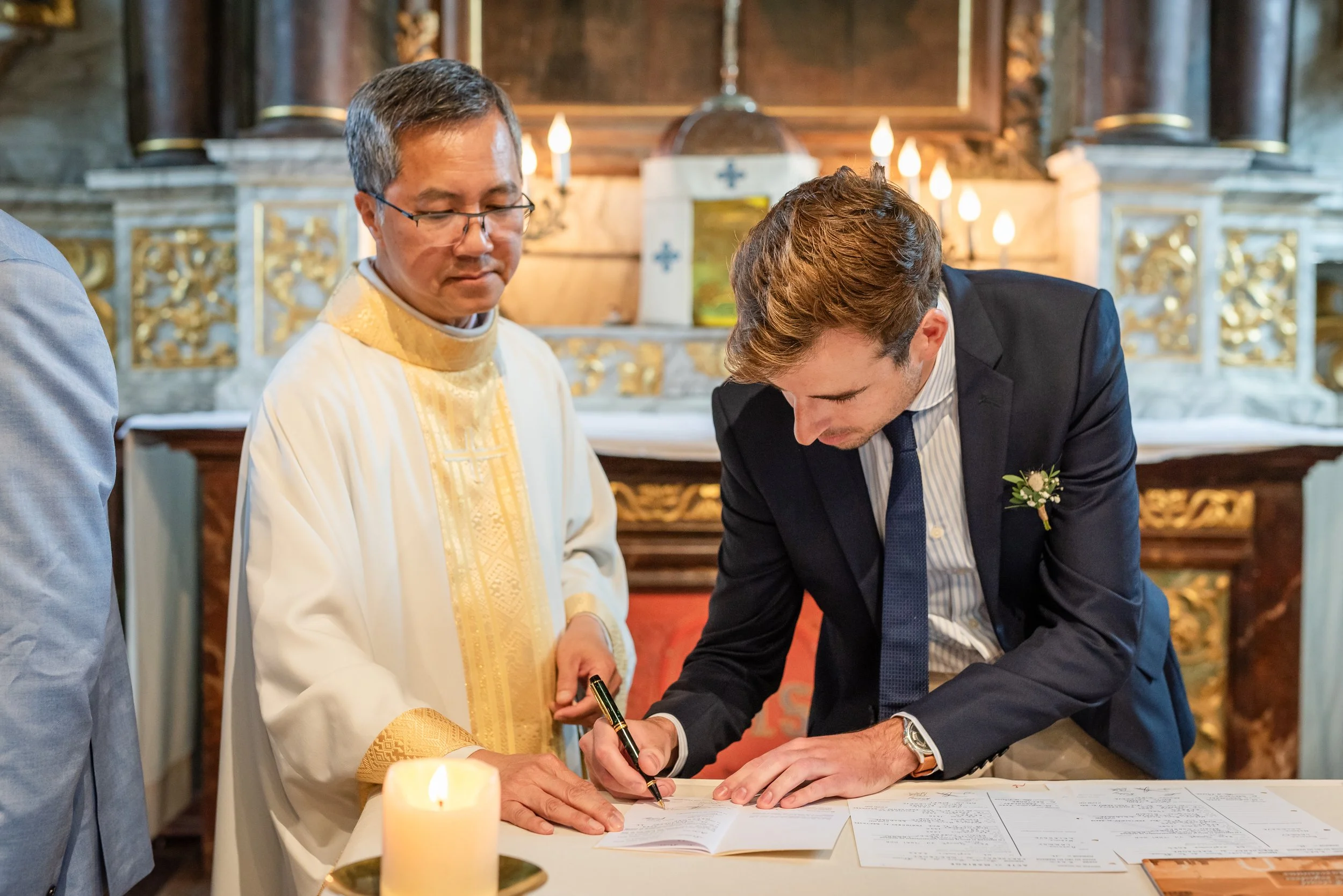 Un homme en costume noir signe un document pendant qu'un prêtre en vêtements liturgiques assiste, dans une église ornementée, avec une bougie allumée au premier plan.