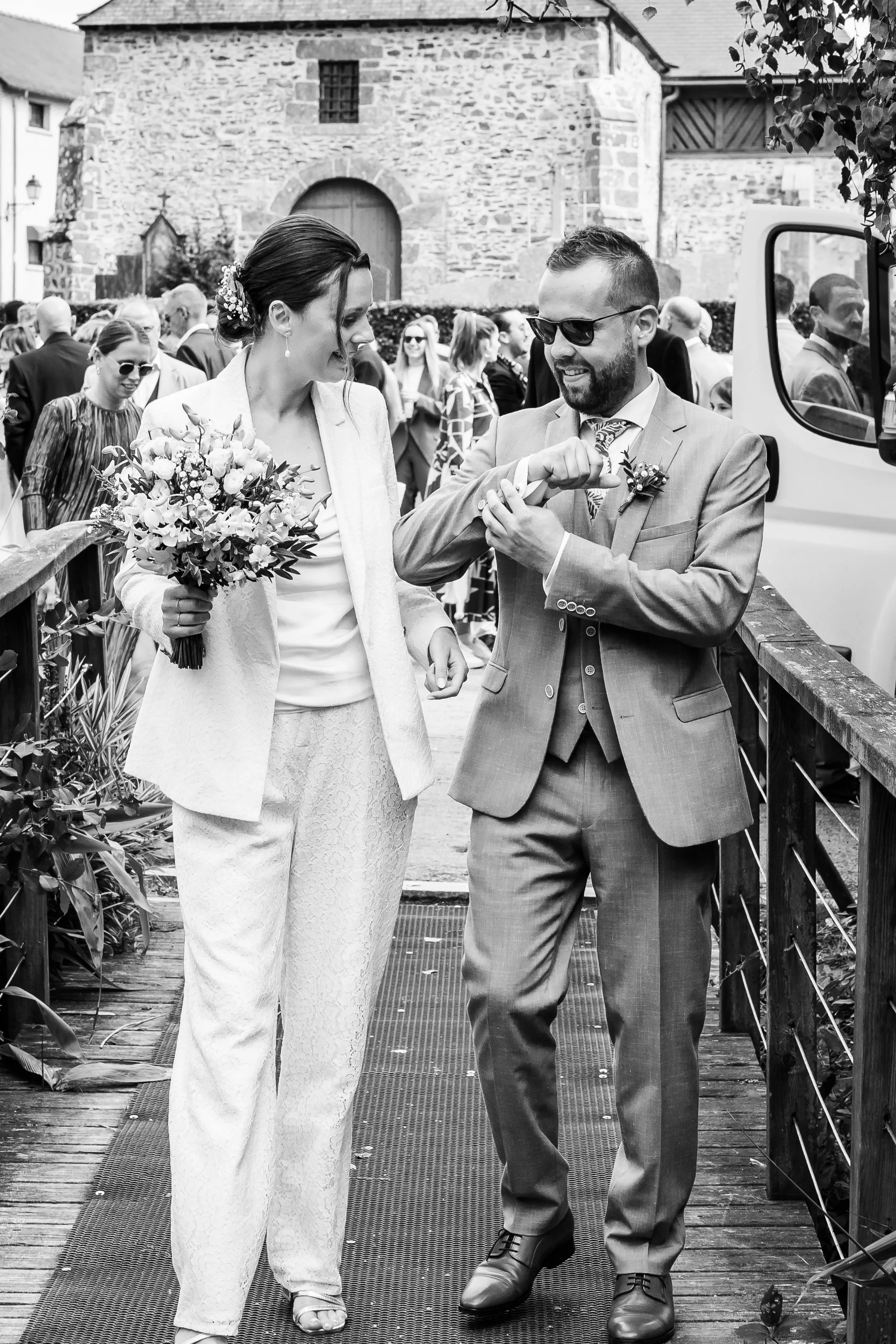 Un couple en costume de mariage échangeant des bagues lors d'une cérémonie en plein air, avec un groupe de personnes en arrière-plan.
