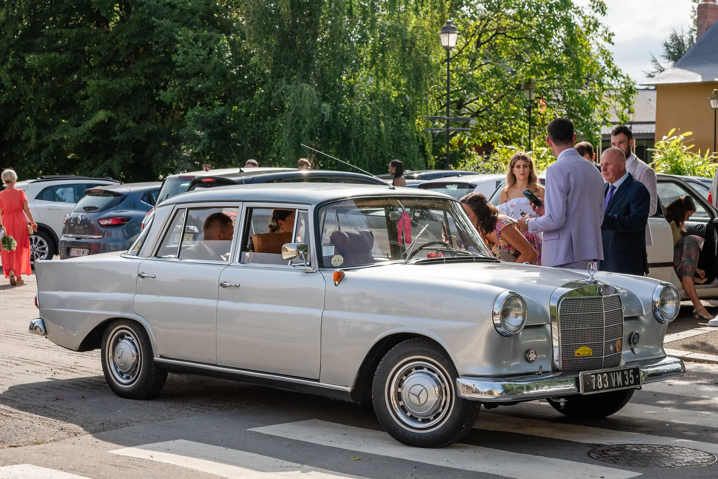 Une voiture ancienne blanche Mercedes-Benz stationnée dans une cour en compagnie de plusieurs personnes, dont un homme en costume bleu et une femme en robe blanche, avec des arbres verts en arrière-plan.