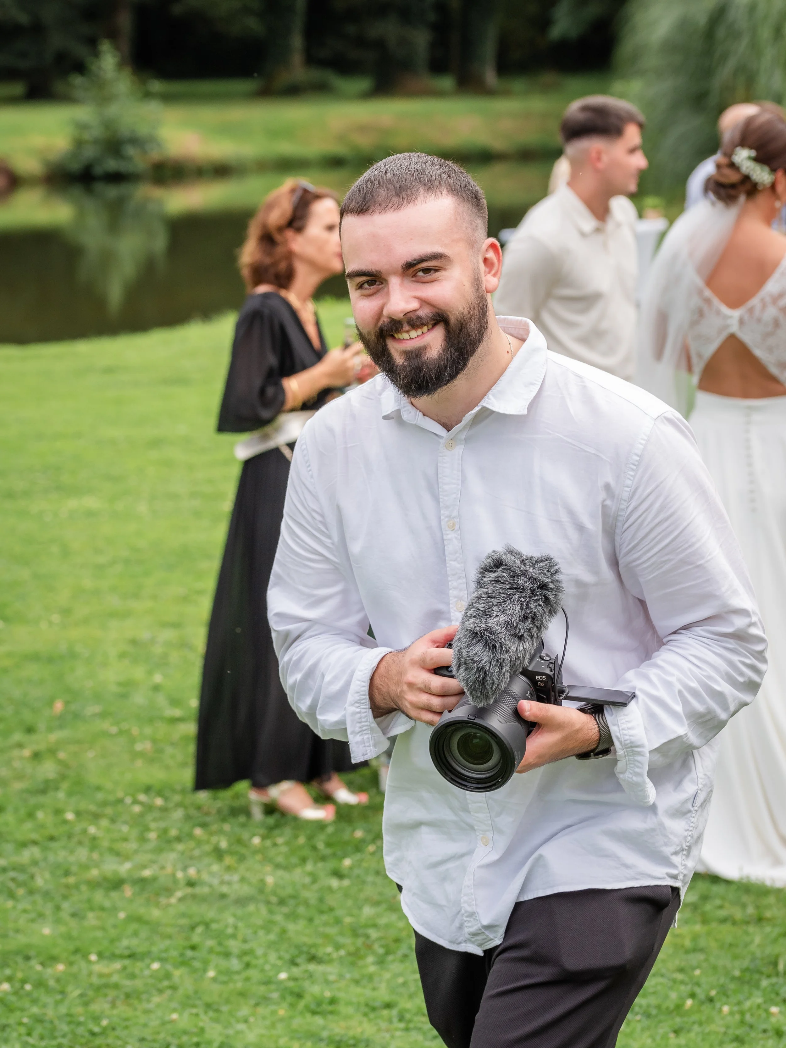 Homme souriant tenant un appareil photo avec micro et porte une chemise blanche, assiste à un mariage dans un jardin avec des invités en arrière-plan.