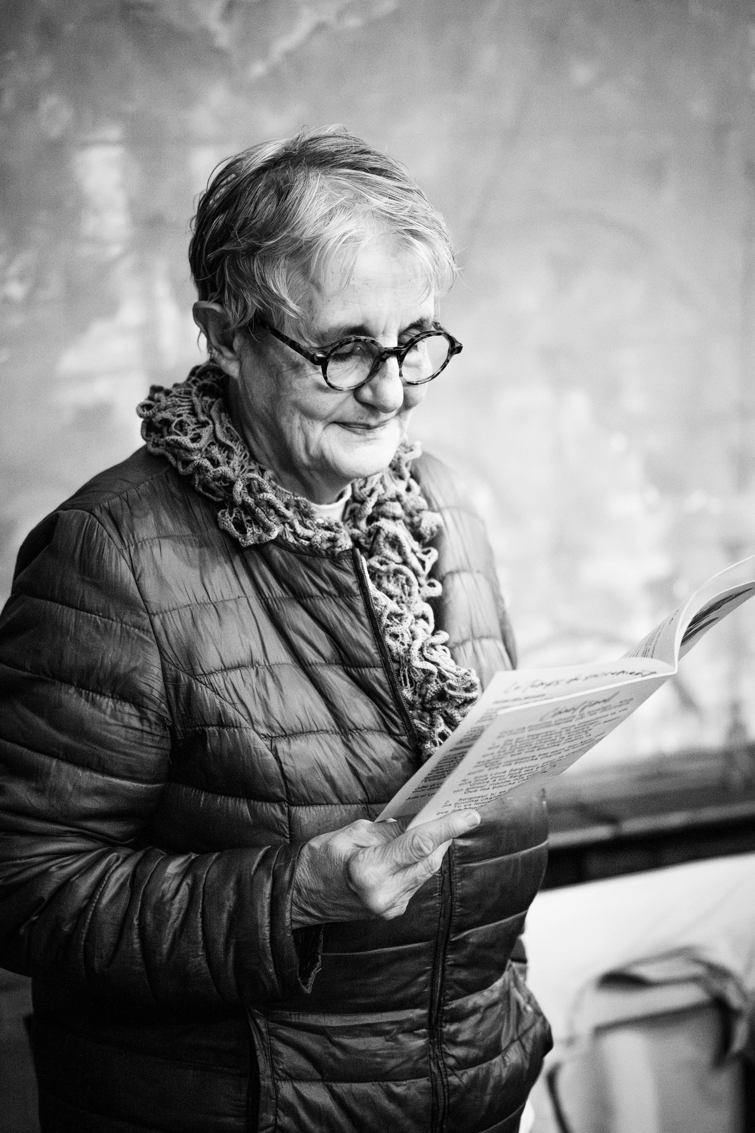 Une femme âgée portant des lunettes, vêtue d'une veste matelassée et d'une écharpe en crochet, lit un document ou une lettre.